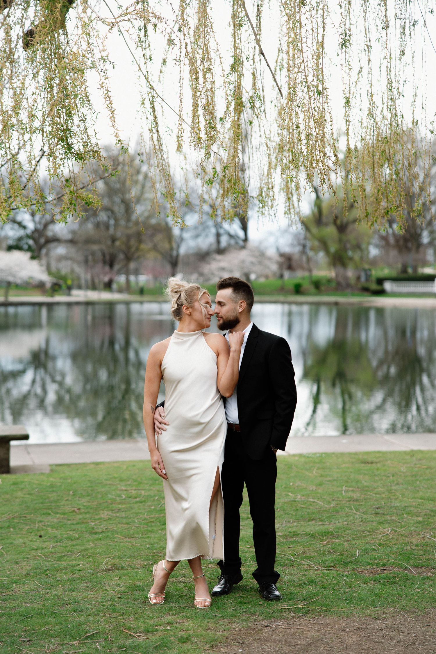 Couple in formal attire shares intimate moment by reflective pond under weeping willow trees. Cleveland, OH