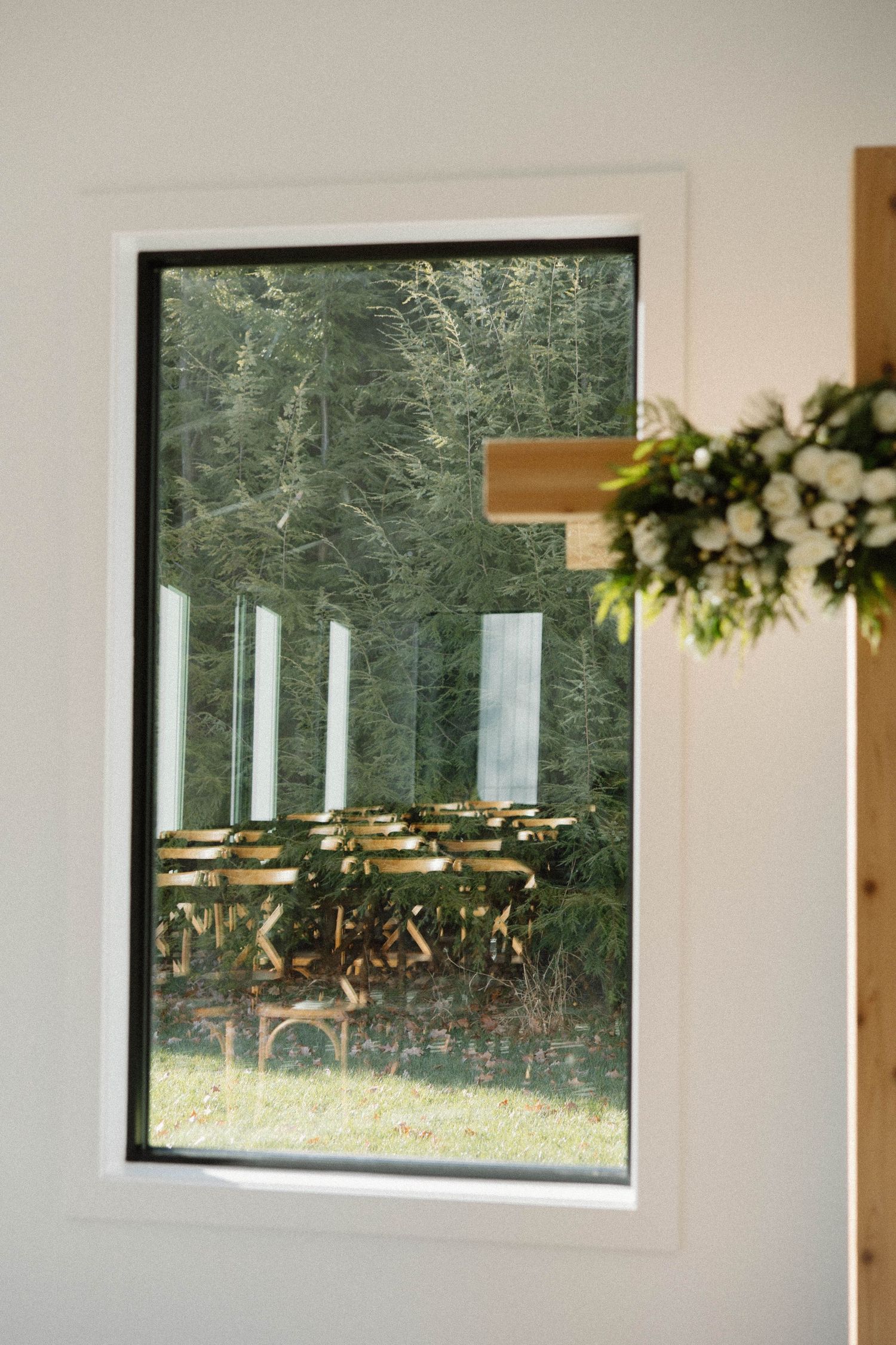 Window frame view of wooden chairs arranged outdoors with evergreen trees in background.