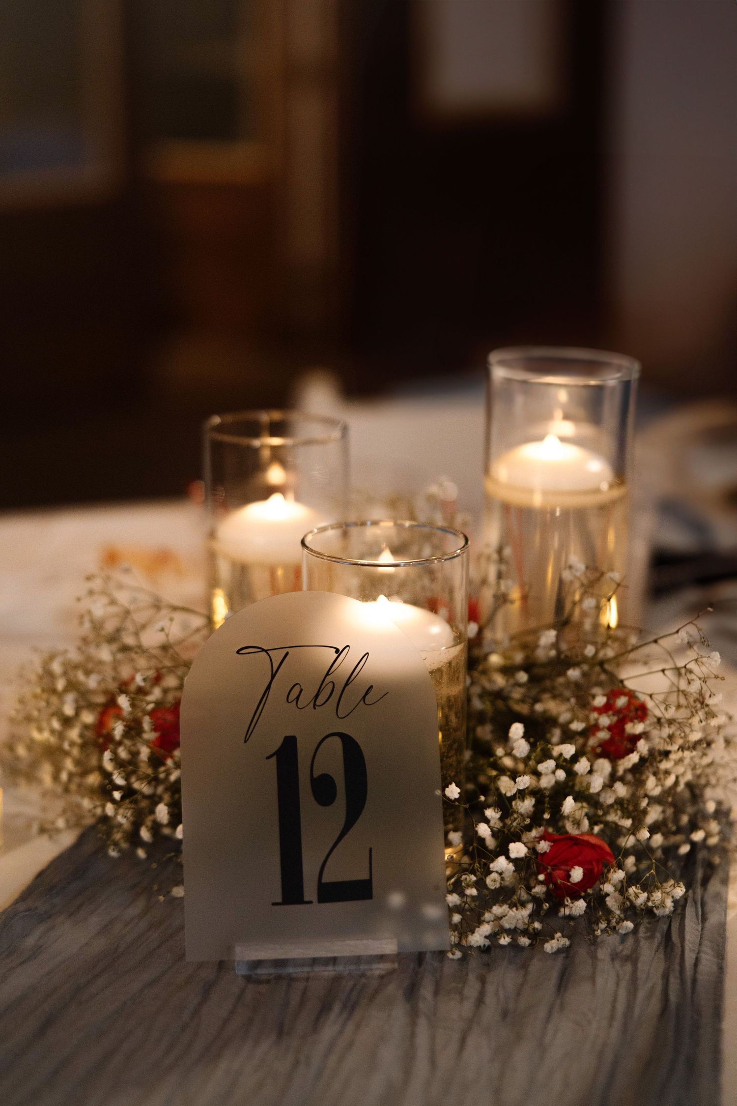 Wedding table number 12 displayed in a glass votive holder surrounded by lit candles and baby's breath floral accents.