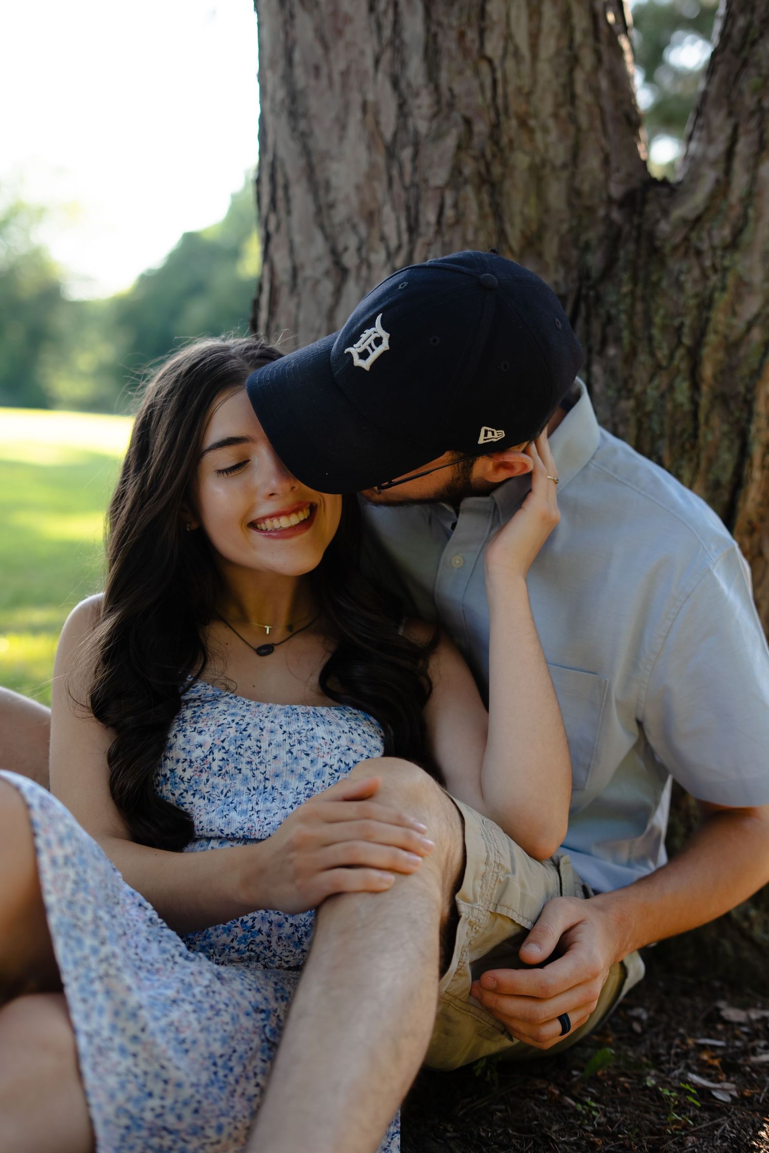 A couple sits together under a large tree on a sunny day, with one wearing a Detroit baseball cap and the other in a floral dress.