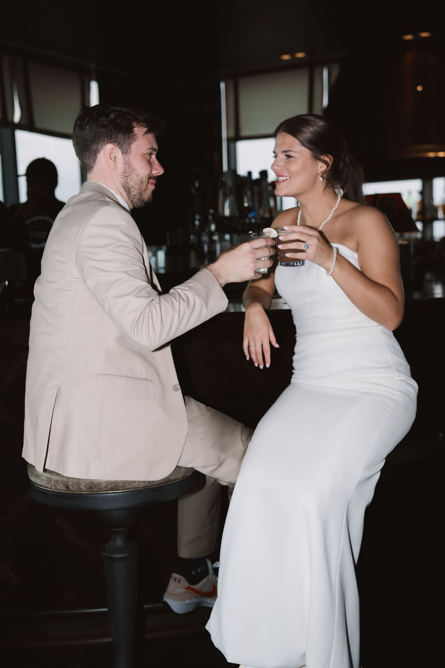A couple in wedding attire shares a toast while sitting at a dark bar.