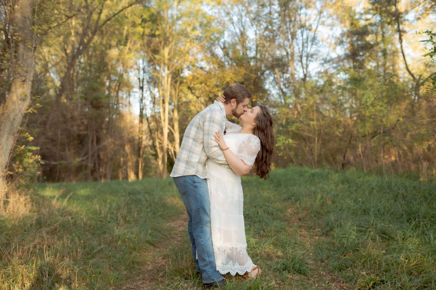 A romantic couple embraces in a golden-lit forest setting during sunset, with the woman wearing a white lace dress.