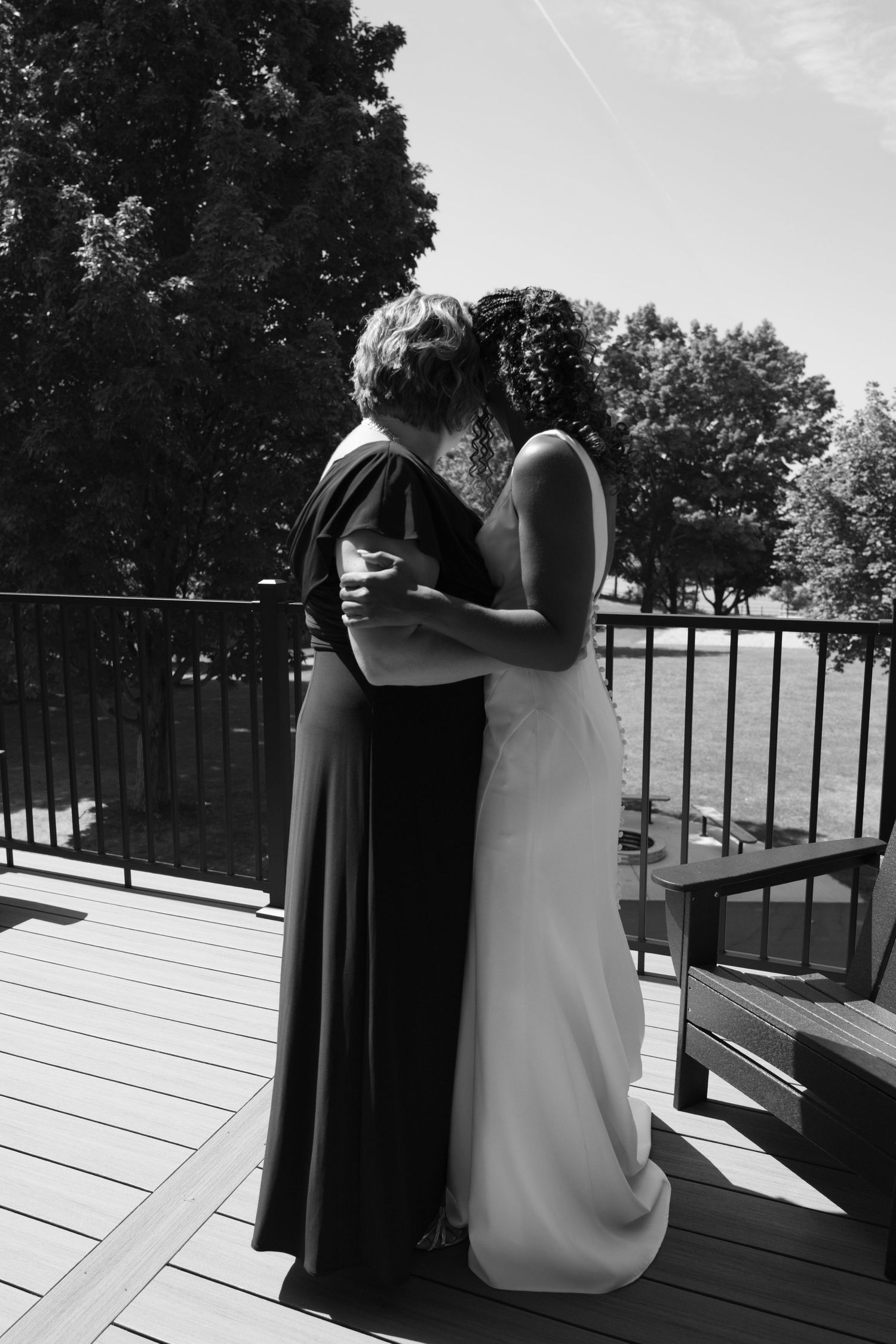 Two people share an intimate embrace on a wooden deck with trees visible in the background, captured in black and white.