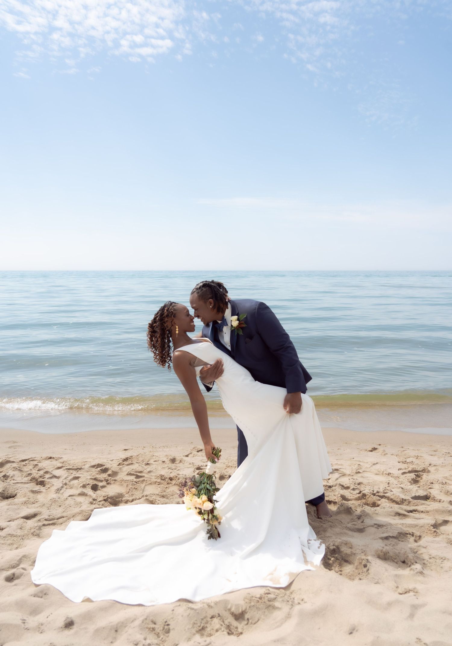 Wedding couple shares romantic kiss on sandy beach with ocean waves in background.
