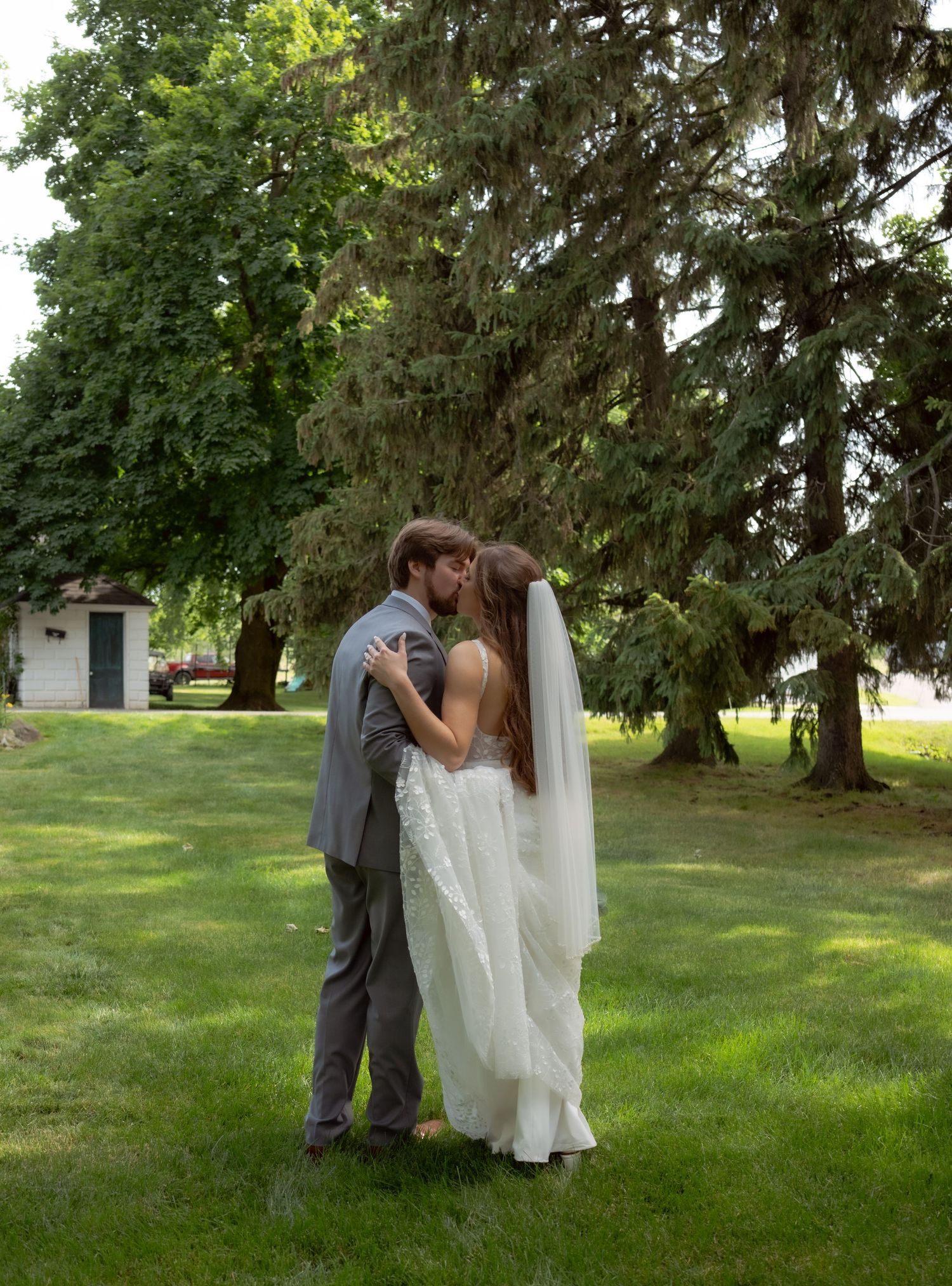 A romantic wedding moment captured outdoors on a lush green lawn with tall evergreen trees in the background.