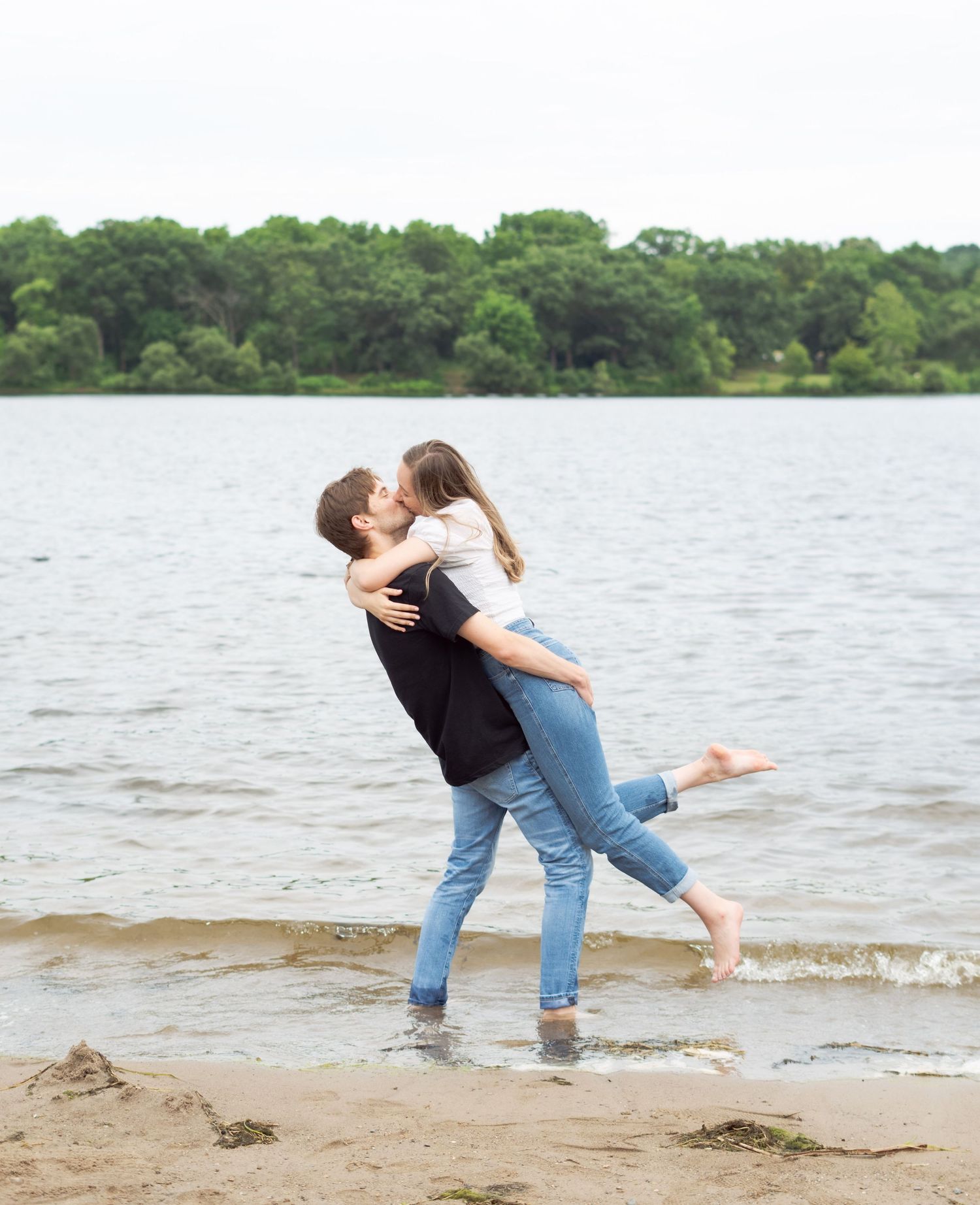 Couple embracing and kissing on a beach with a serene lake and forest backdrop.