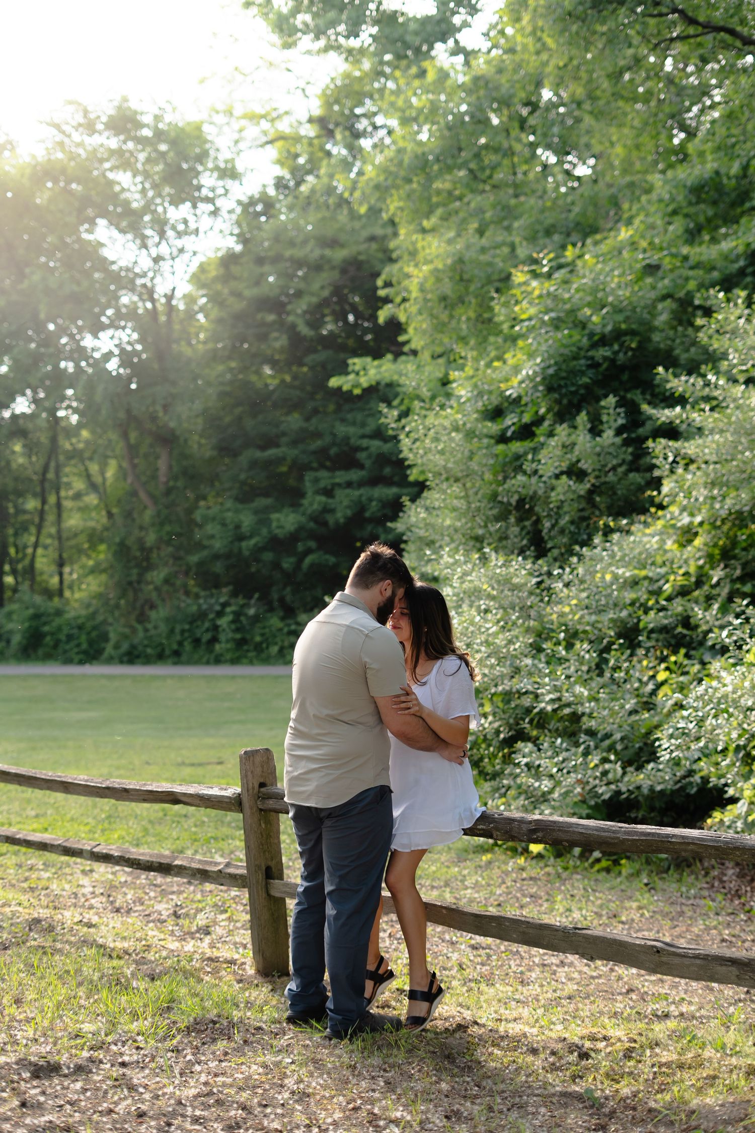 Couple sharing a romantic moment by a wooden fence in a sunlit park during golden hour.