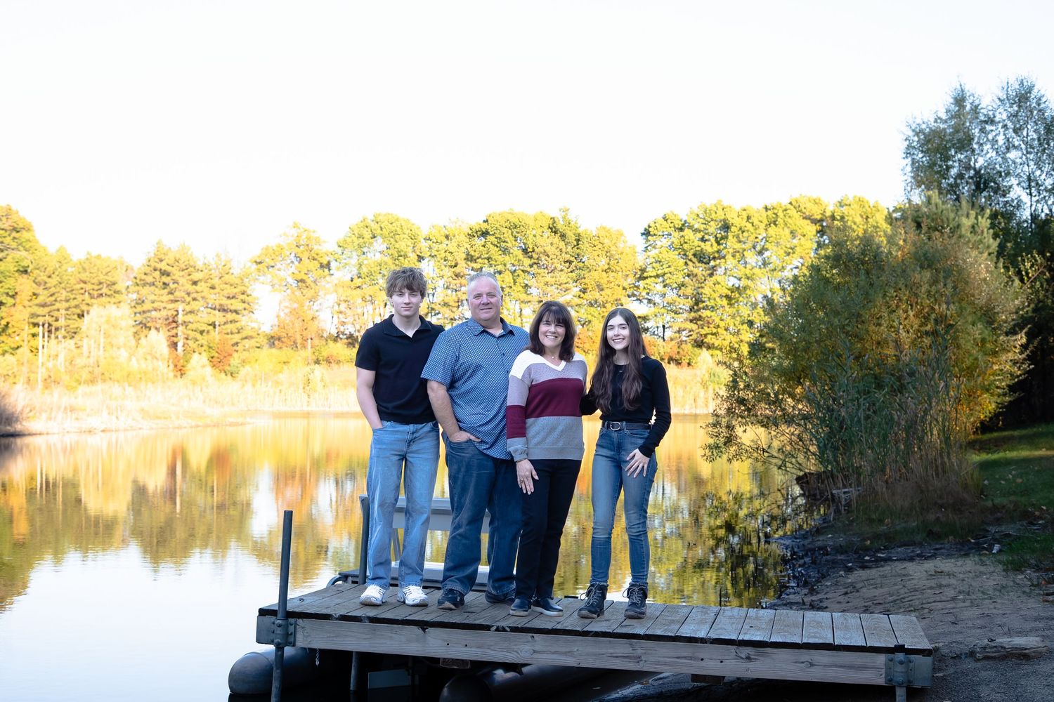 People gathered on a wooden dock by a serene lake during golden hour with trees in the distance.