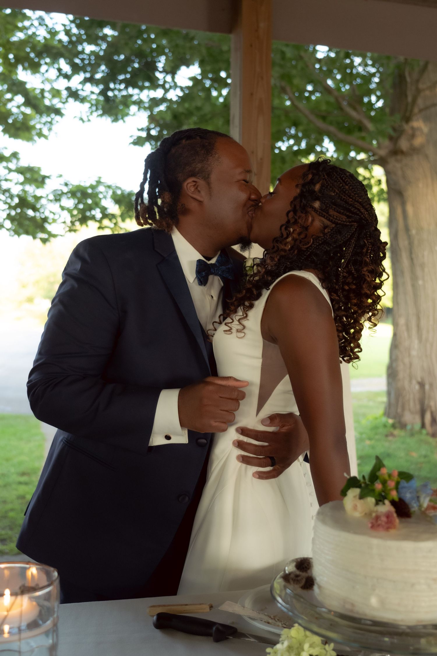 A couple shares a kiss while standing next to their elegant white wedding cake at an outdoor venue.