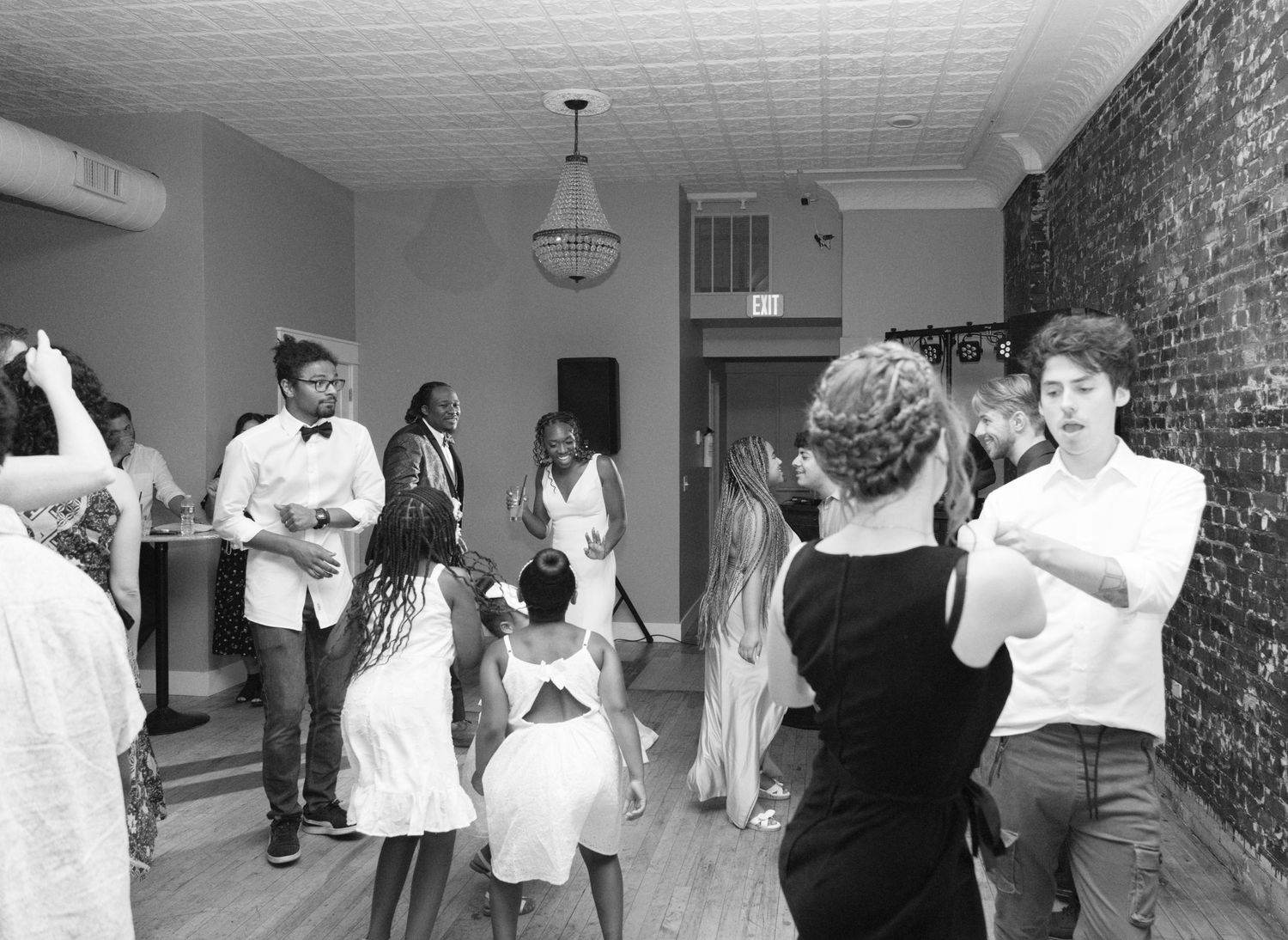 Black and white photograph of a casual party scene in a hallway with exposed brick walls and a hanging light fixture.