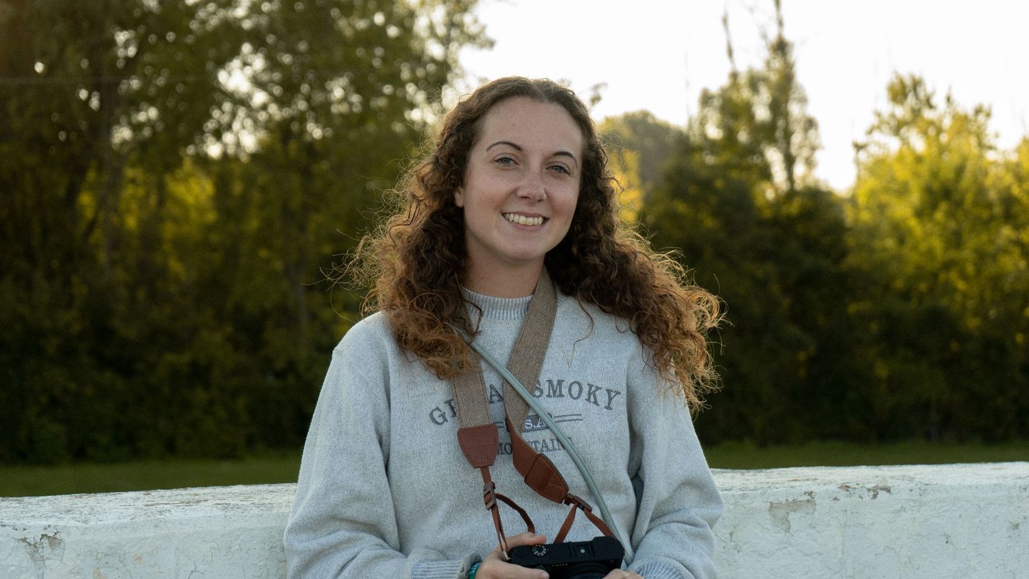 A person in a light gray sweatshirt with curly hair sits outdoors on a sunny autumn day.