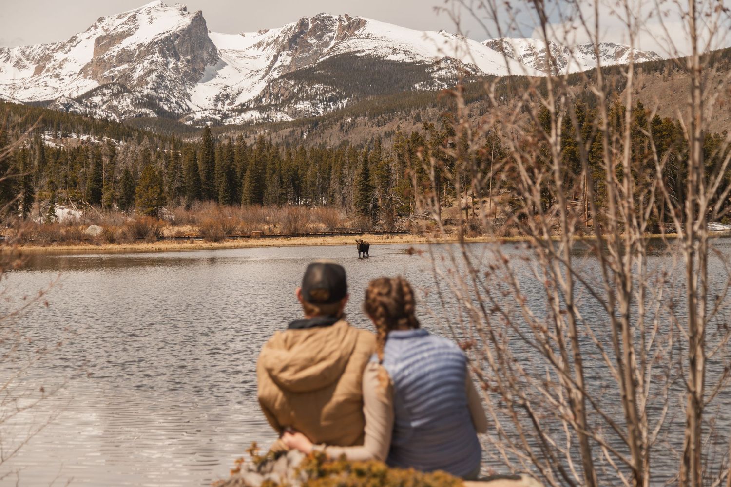 Two people admiring a scenic mountain lake view with snow-capped peaks and forest landscape in the background.