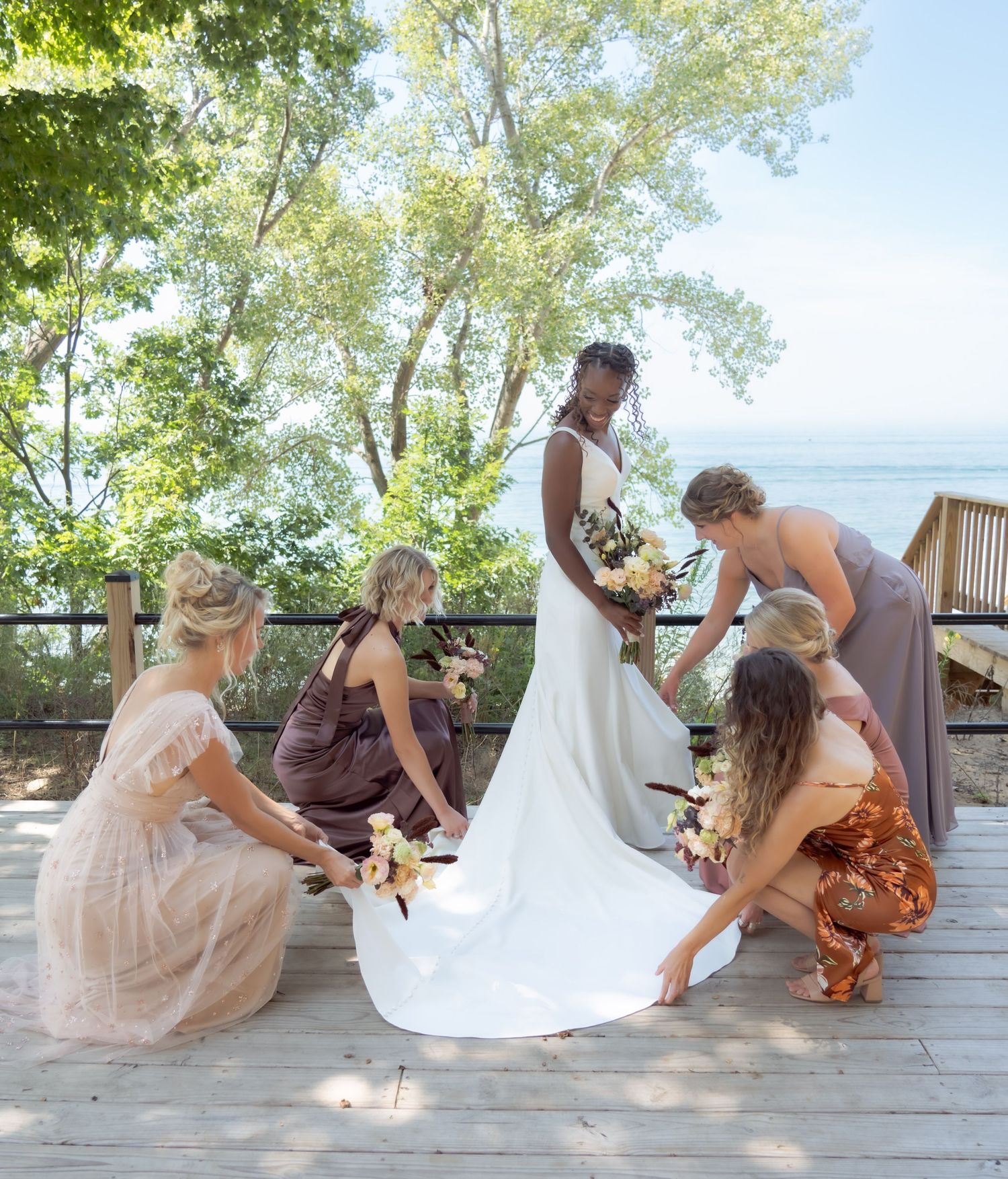 Wedding party helping adjust bride's train on outdoor deck with lake view.