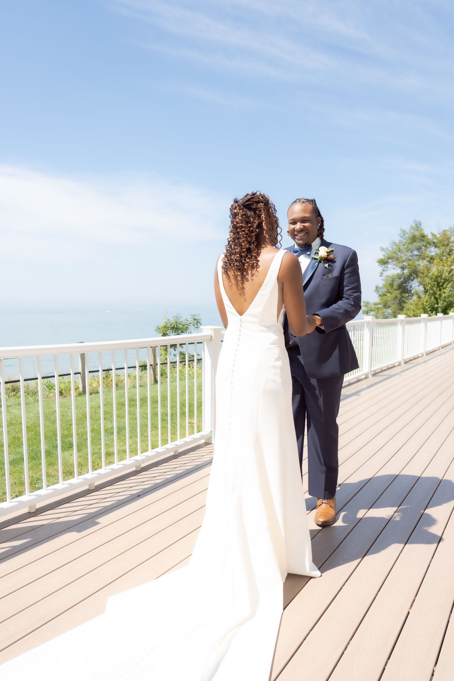 Wedding couple shares first look moment on wooden deck overlooking waterfront on sunny summer day.