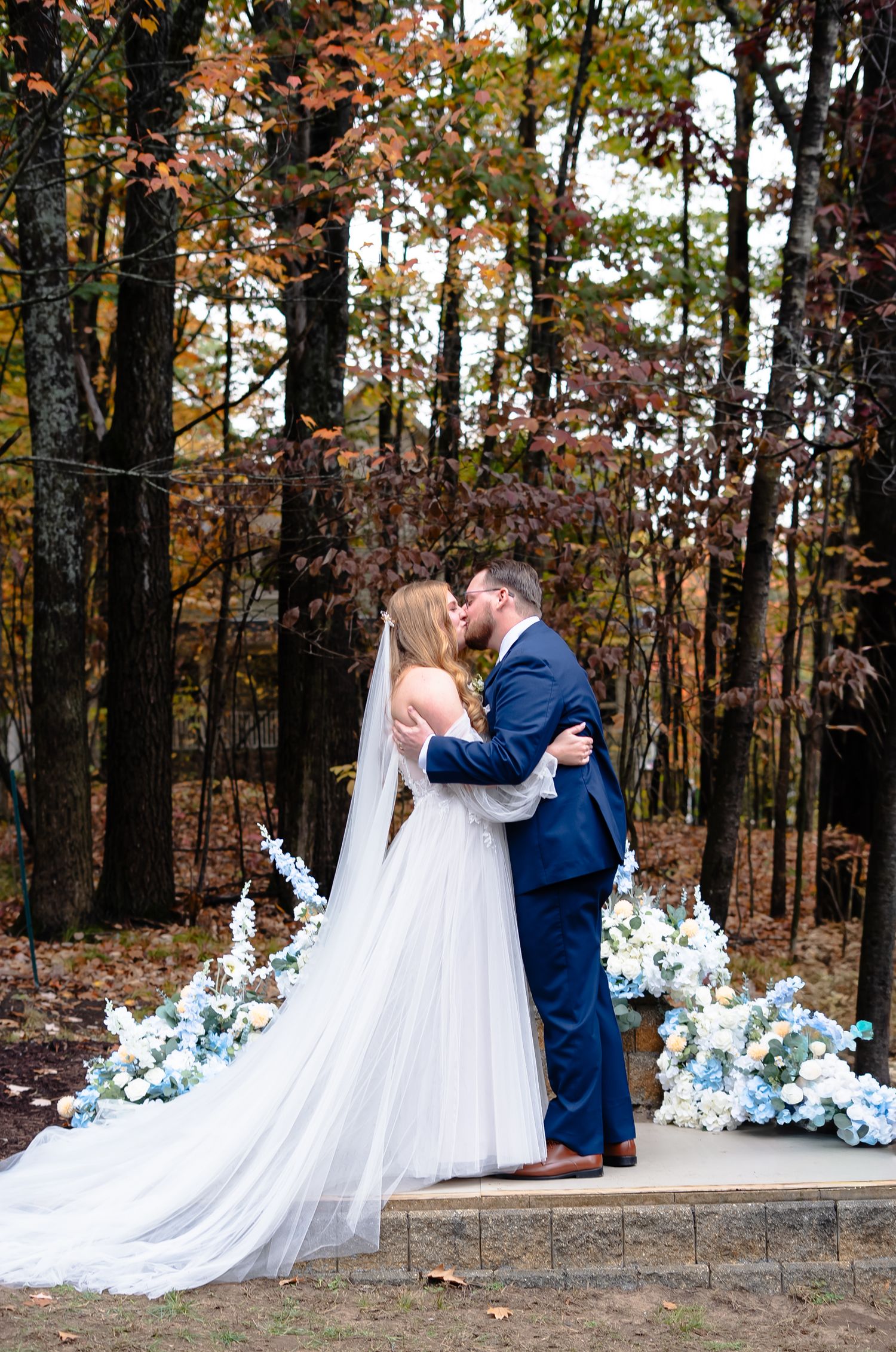 Romantic outdoor wedding couple embraces in front of floral arch with blue and white flowers in autumn forest setting.