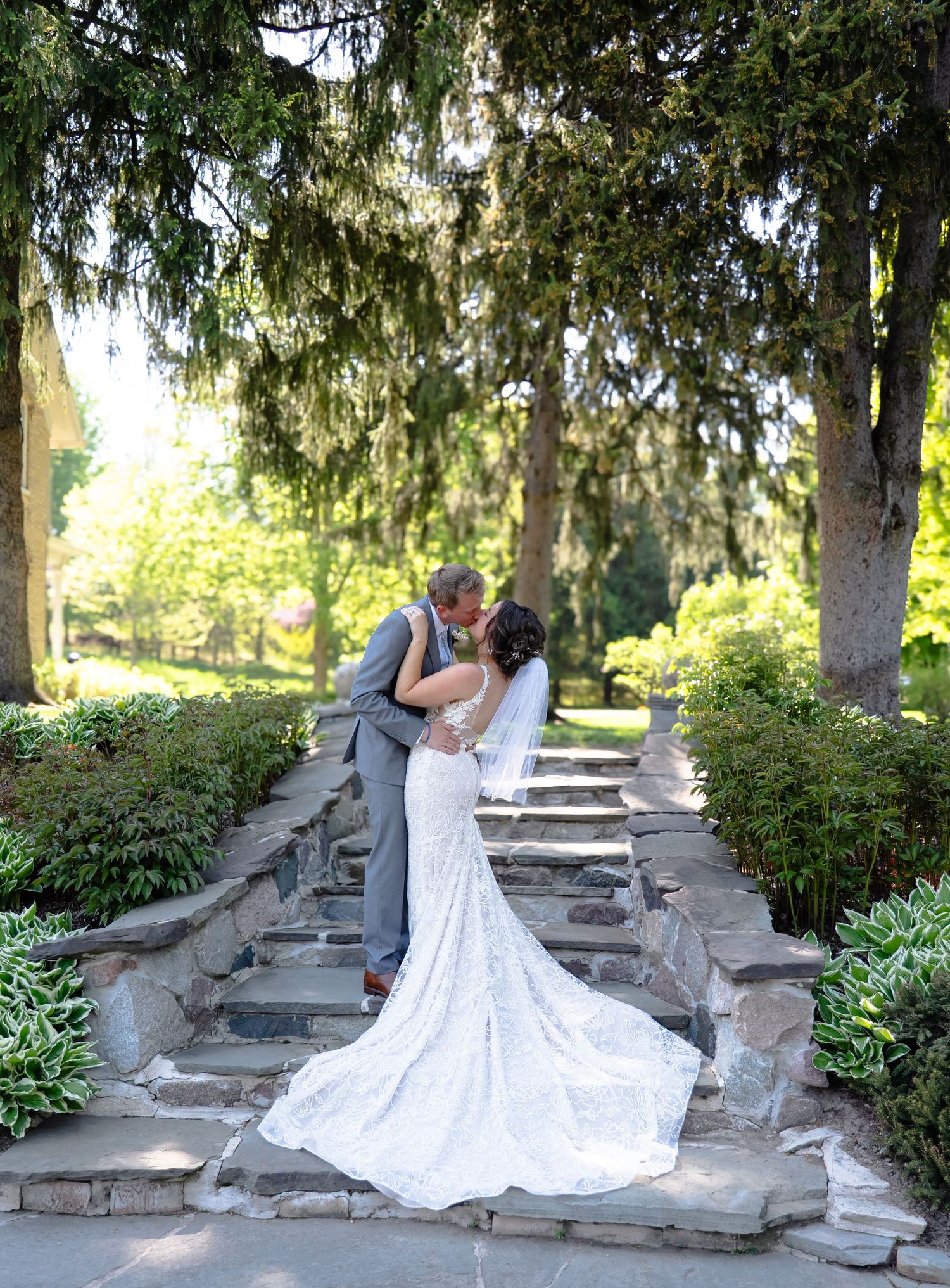 Romantic couple embraces on stone steps surrounded by evergreen trees and lush gardens.