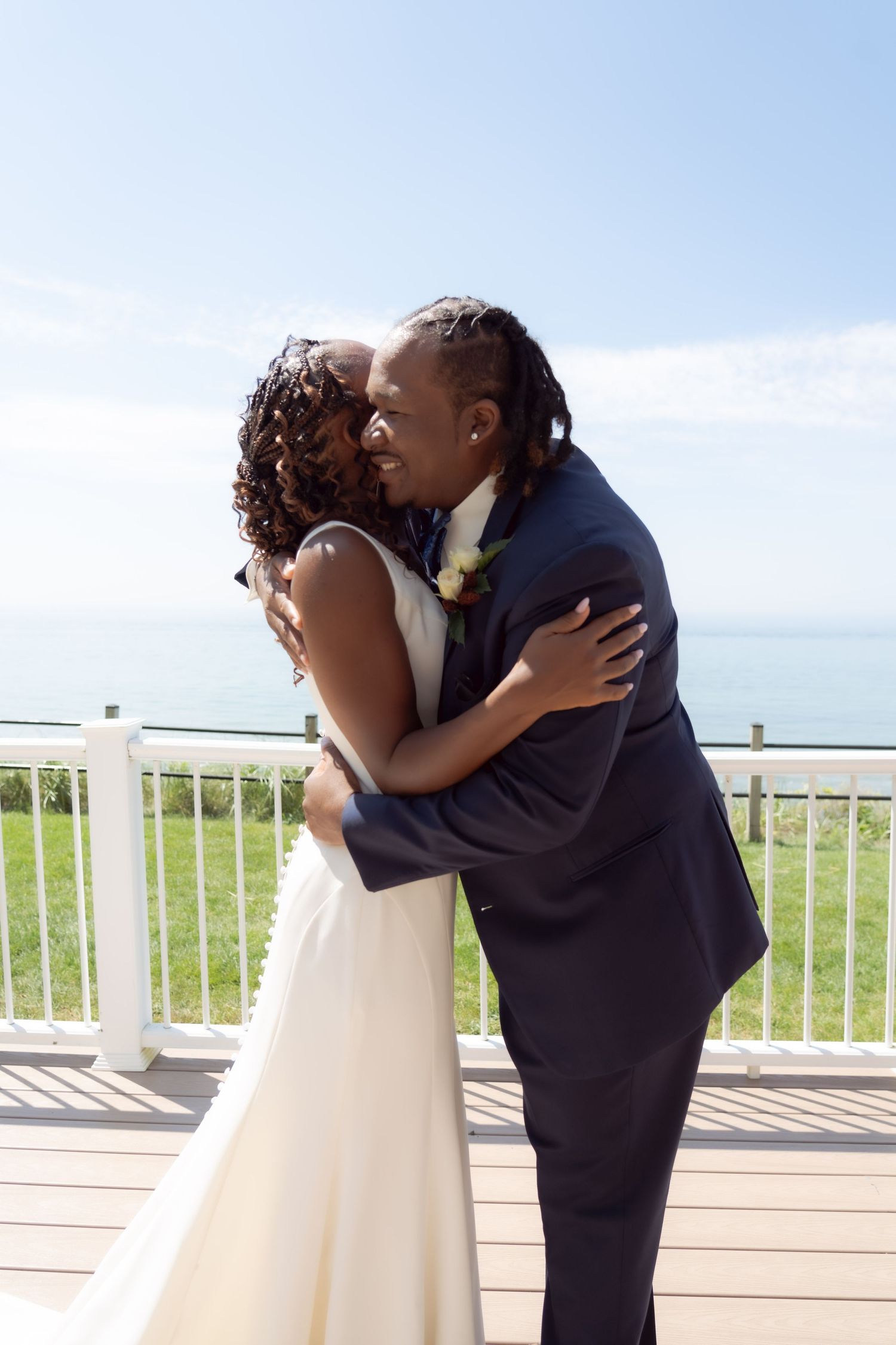 A couple in wedding attire share an intimate moment on a deck overlooking the ocean on their special day.