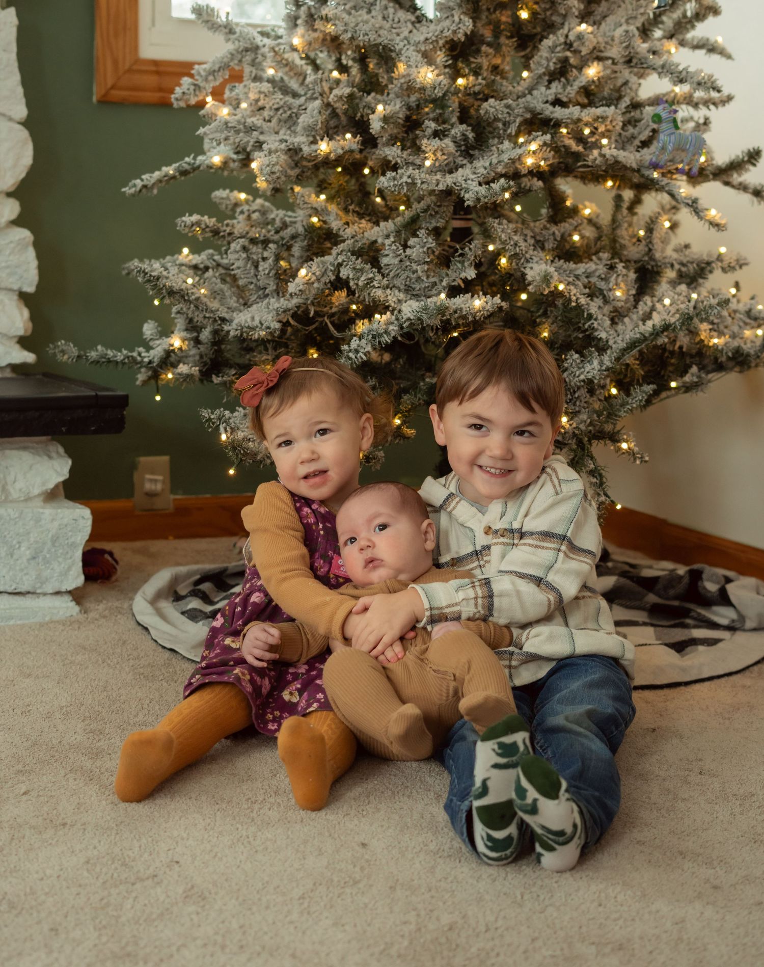 Three children sitting together and smiling in front of a decorated Christmas tree with twinkling lights.