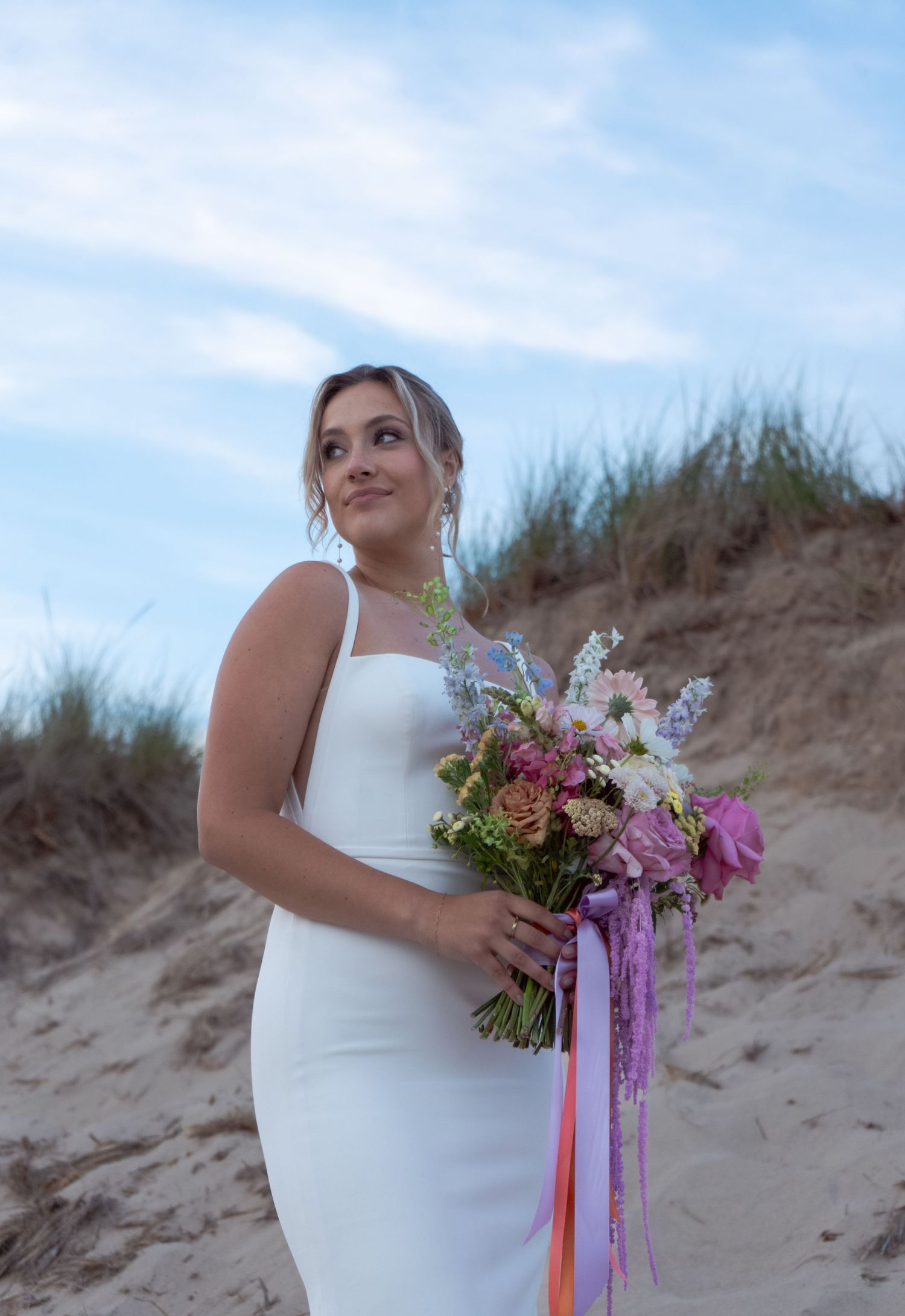 Beautiful bridal bouquet with purple and pink flowers against sandy dunes and blue sky background.