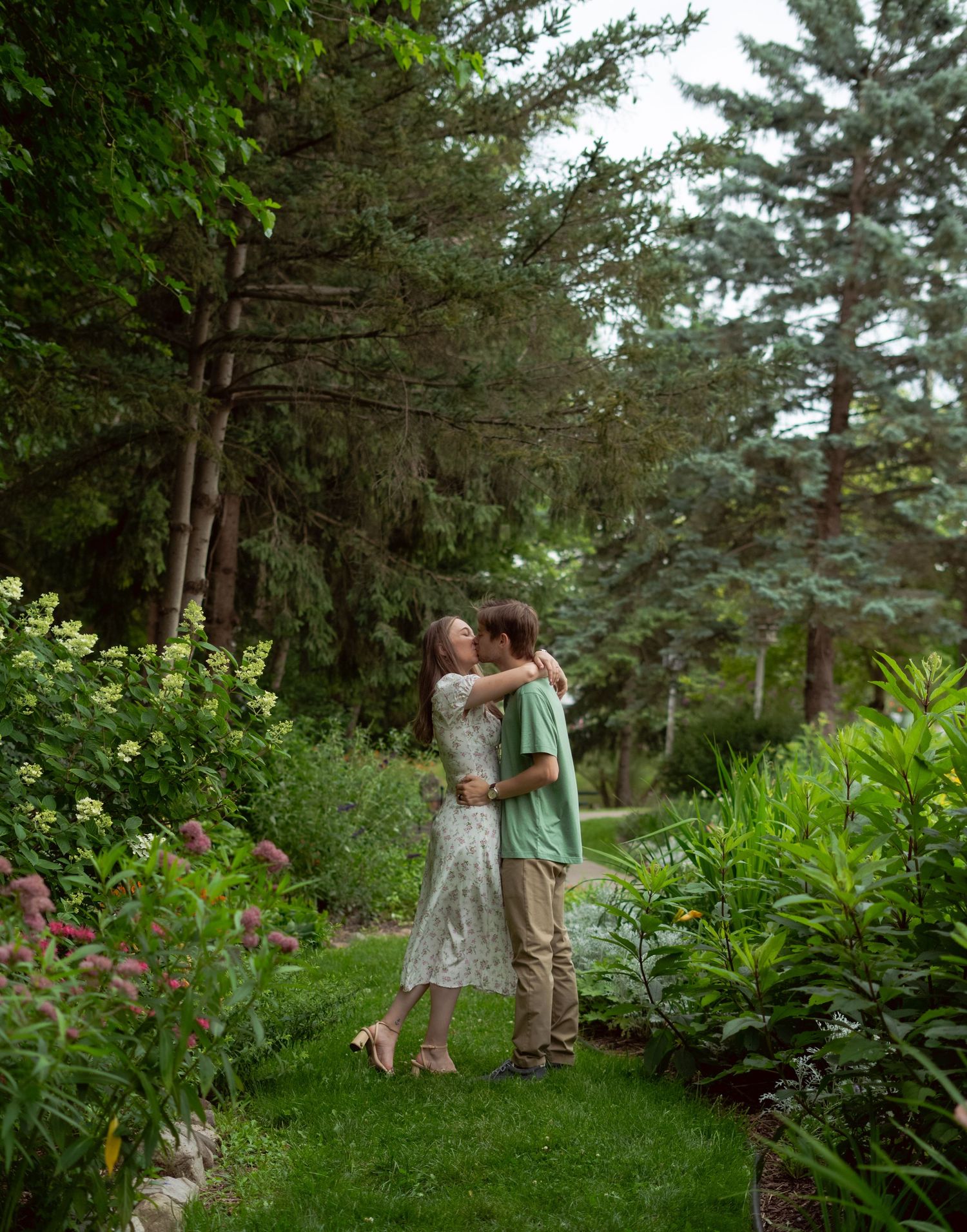 A tender embrace between a couple in a lush garden setting surrounded by evergreen trees.