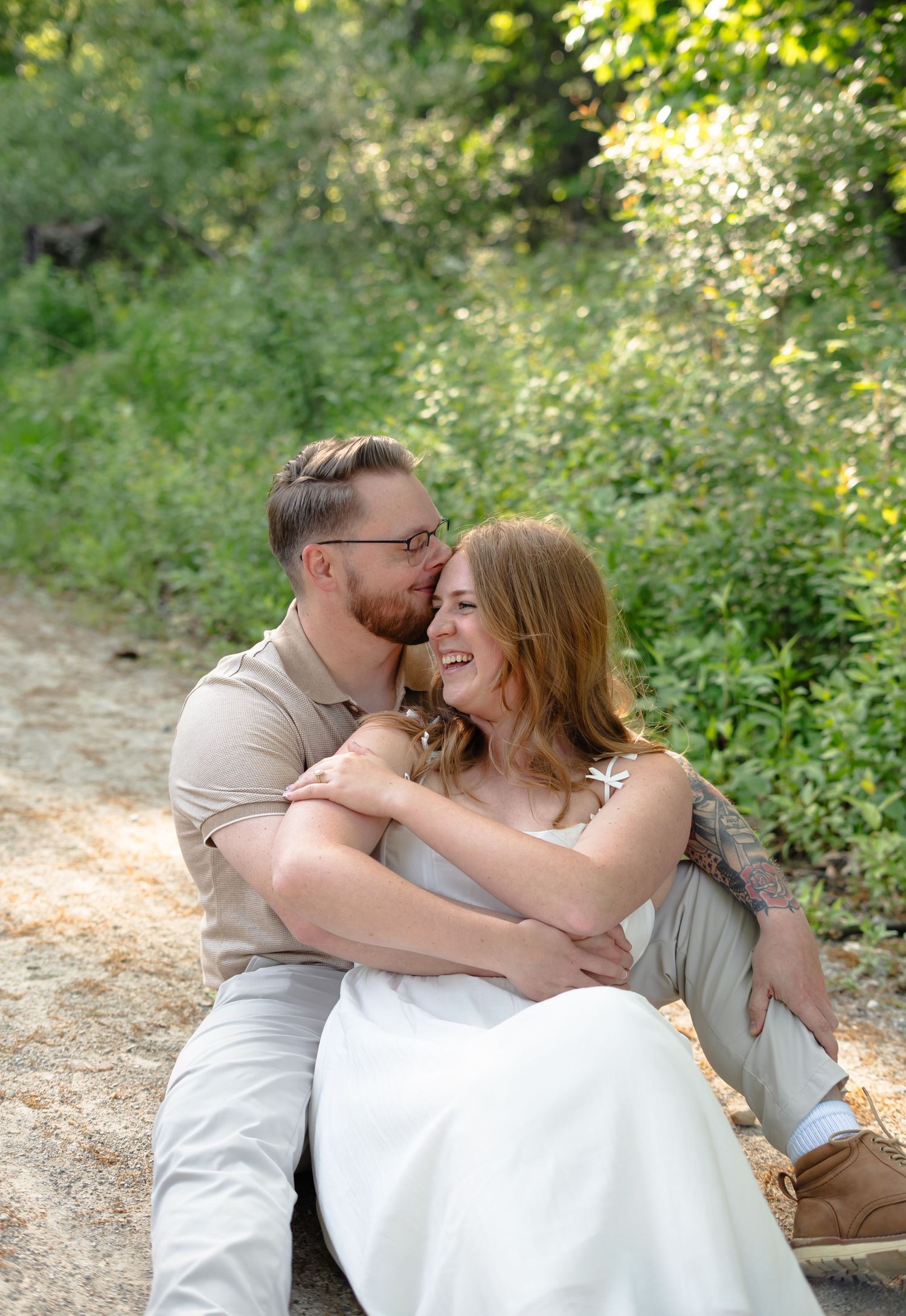 A joyful romantic moment between a couple laughing together while sitting on a sunlit forest path.