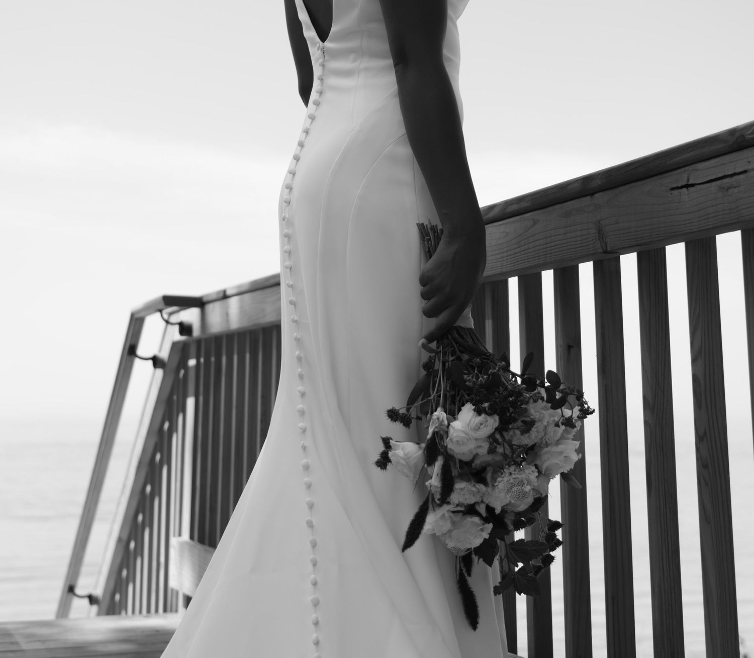 Black and white photograph showing the bottom of a bridal gown and bouquet against a deck railing by the ocean.