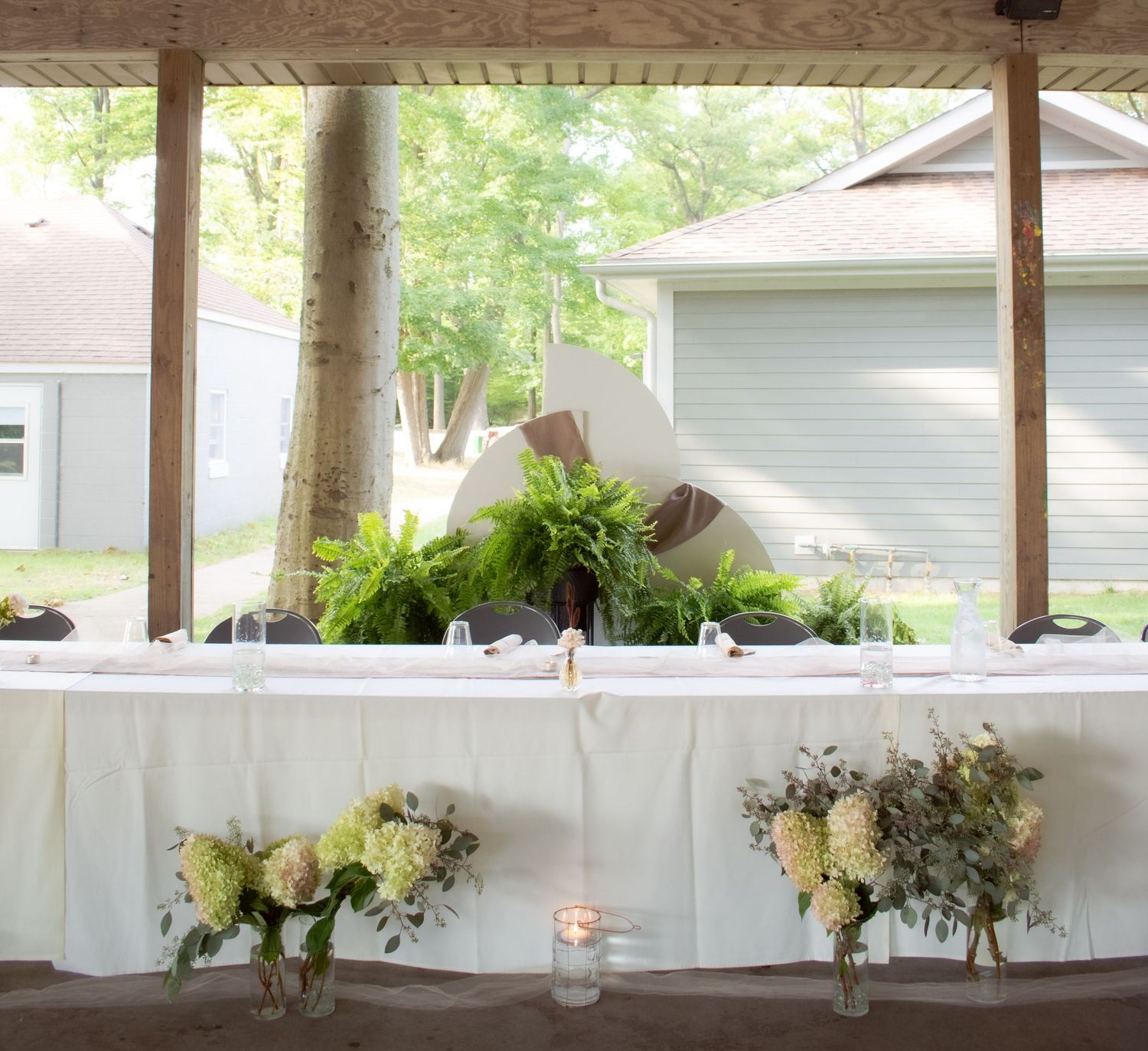 White linen covered table decorated with floral centerpieces and candles for an outdoor reception under a porch.