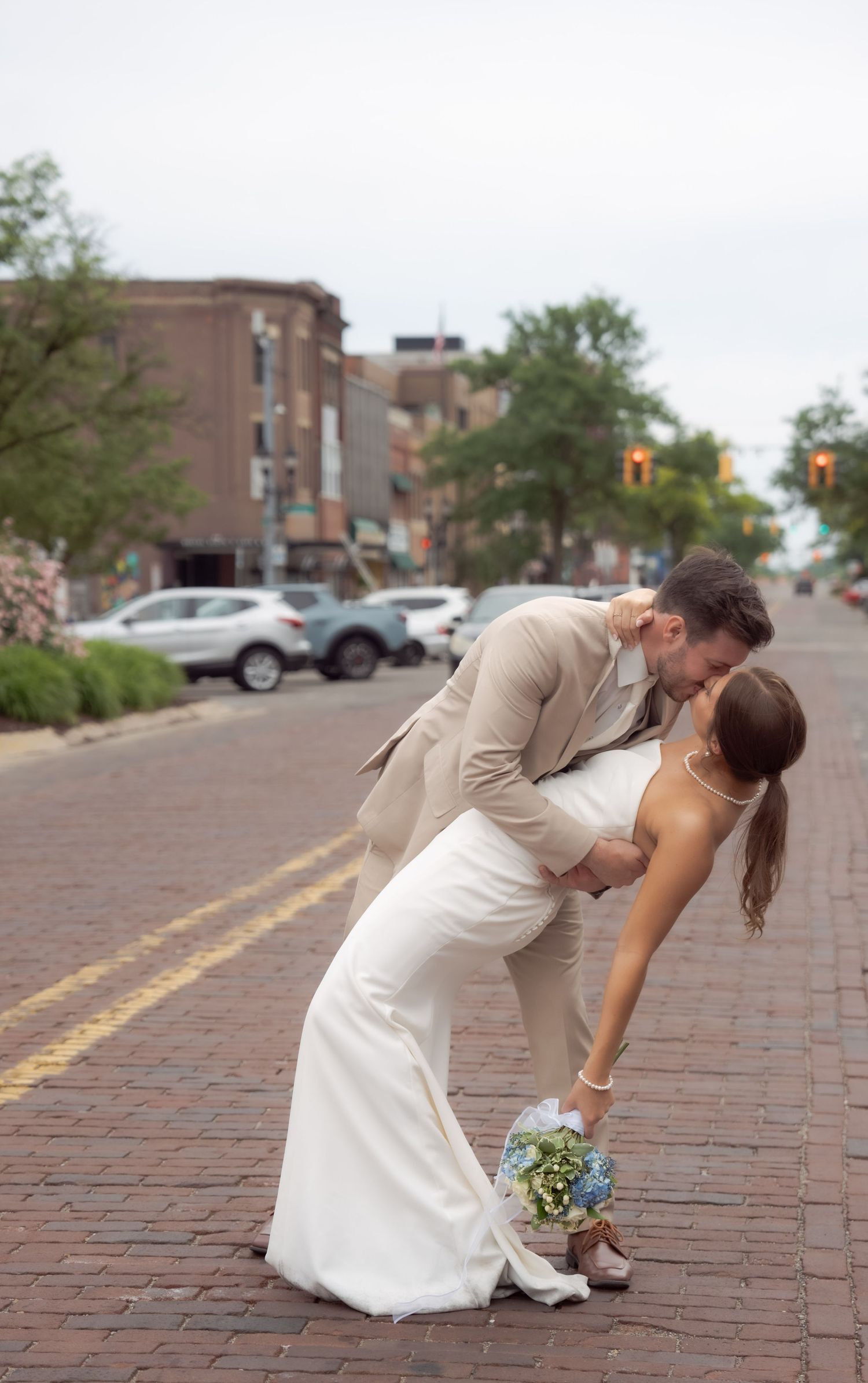 A romantic wedding moment captured on a brick paved street with urban buildings and parked cars in the background.