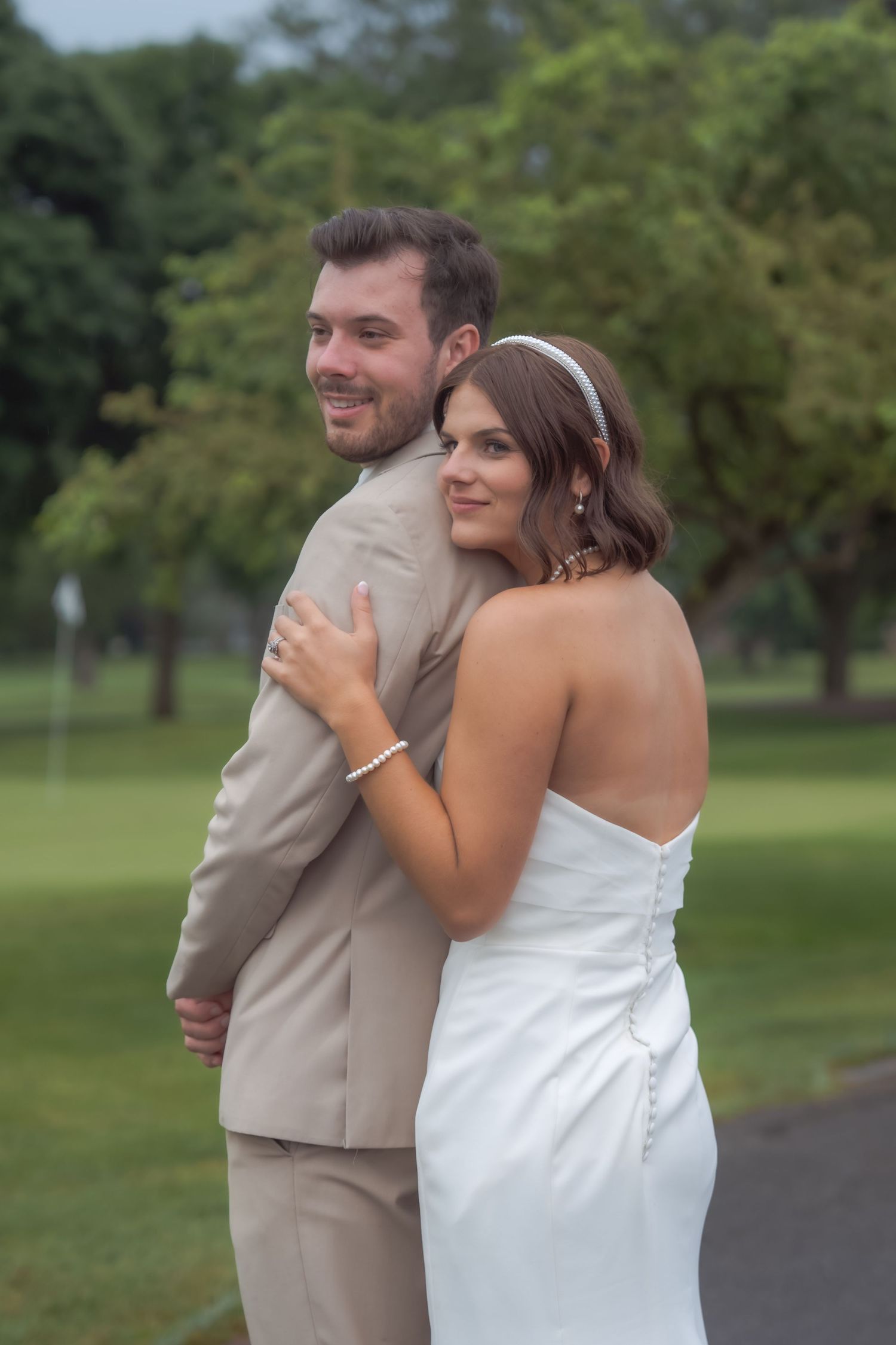A couple poses together outdoors at a wedding, with the bride in white and groom in beige standing on a path near trees.