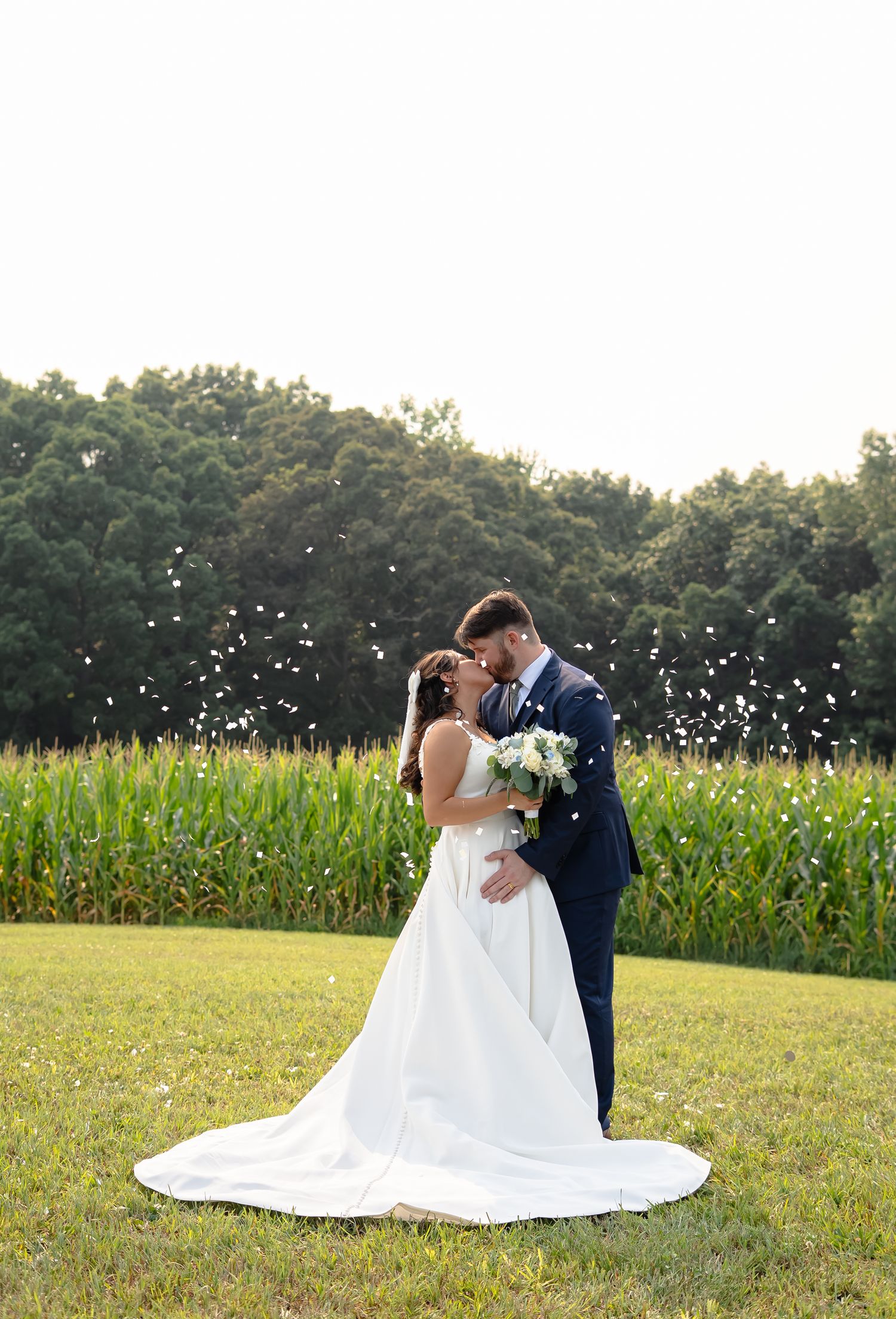 A romantic wedding couple shares intimate moments in a cornfield as white petals float through the summer air.