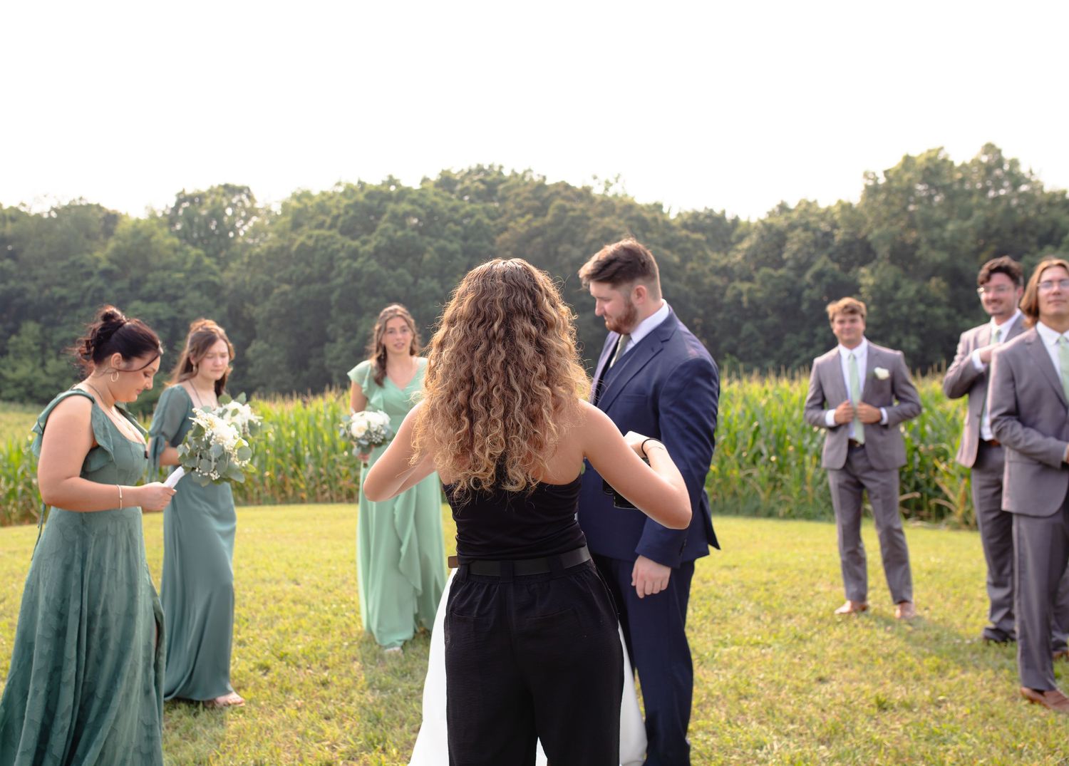 Group of wedding guests in sage green dresses and dark suits gather outdoors in a sunny meadow for photos.