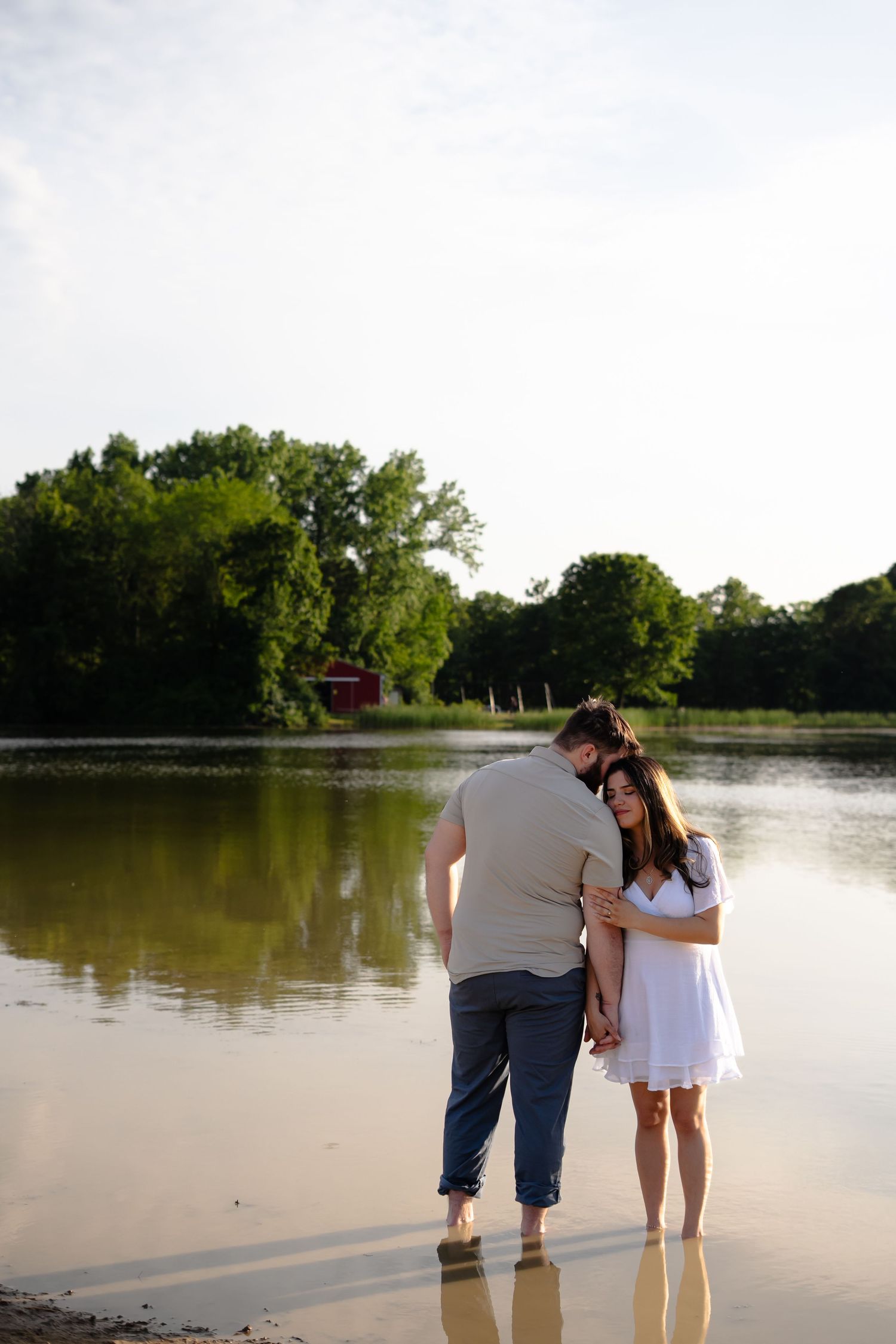 A couple stands together at the edge of a serene lake during golden hour, creating a romantic silhouette against the water.