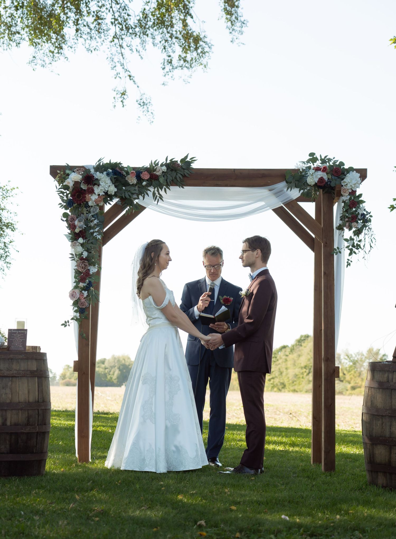 Wedding ceremony under wooden arch decorated with white fabric and floral arrangements.