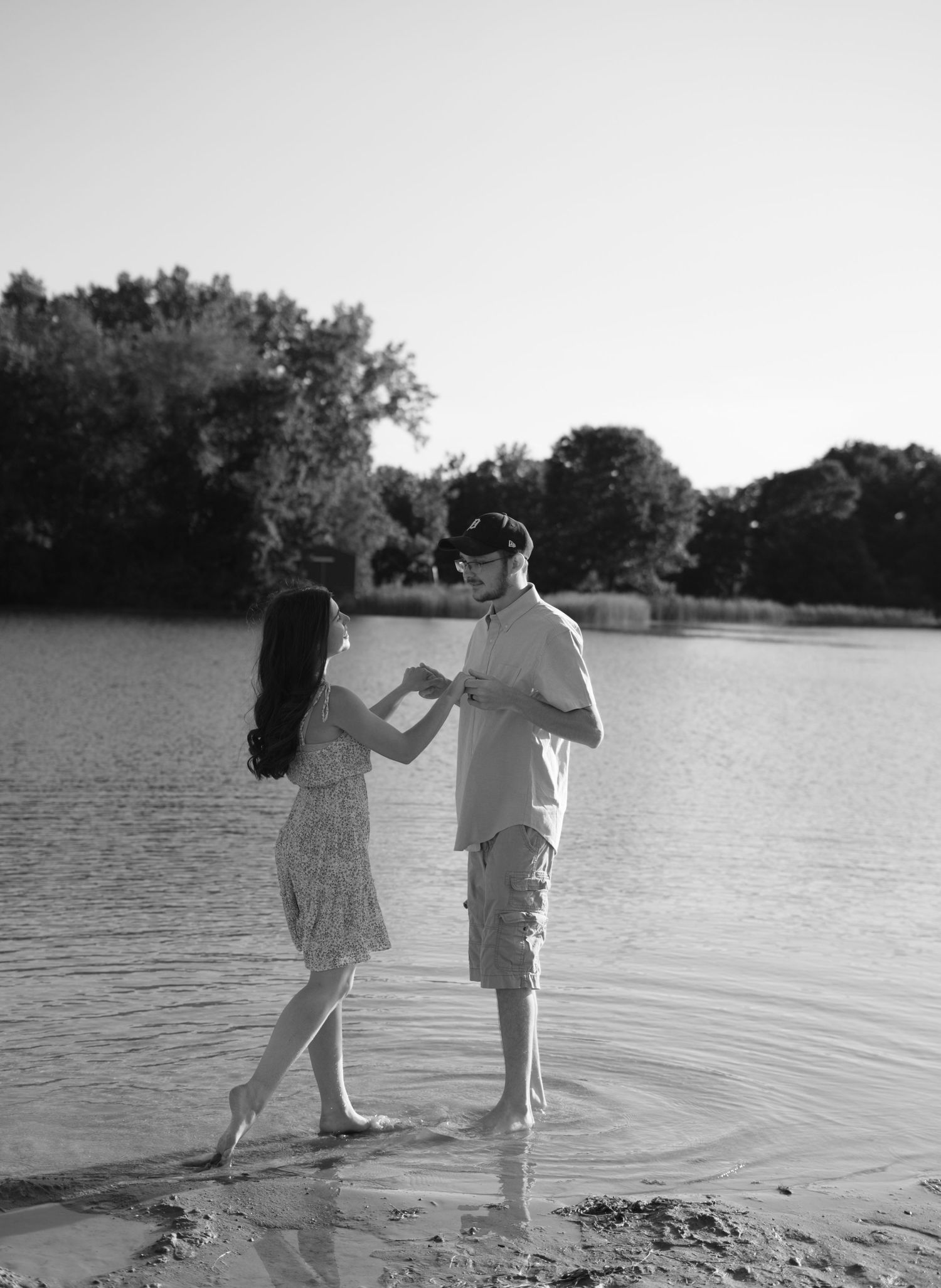 A couple holds hands while dancing in shallow water by the lakeshore at sunset in a romantic black and white scene.