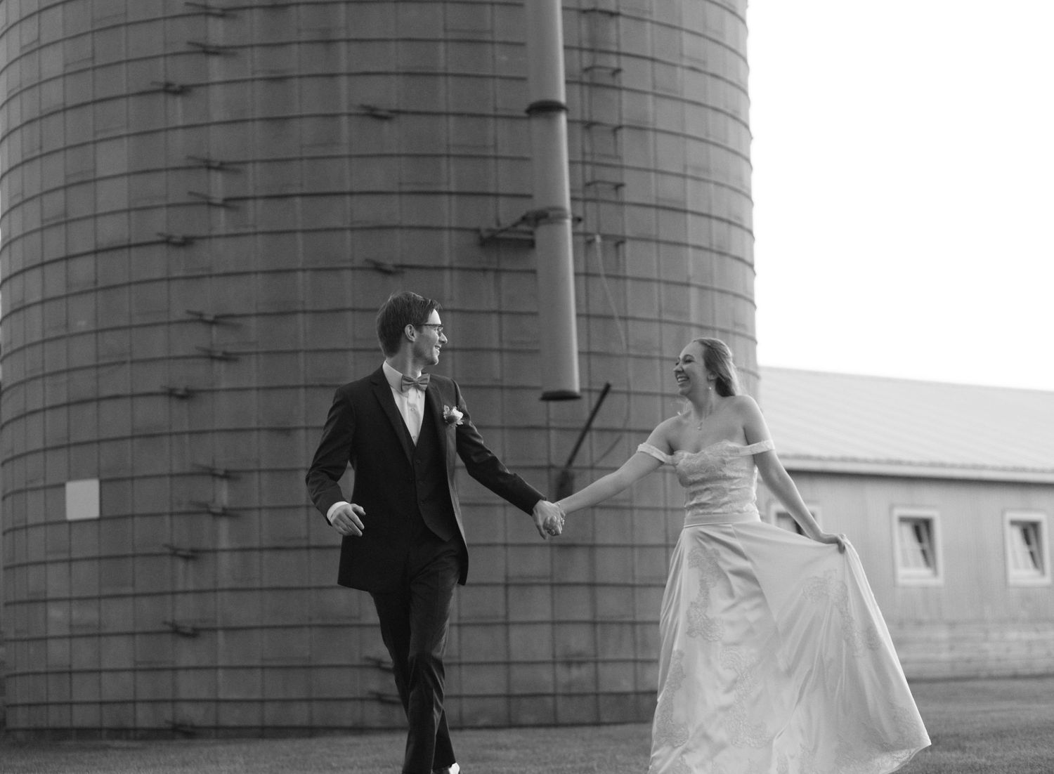 A couple in wedding attire poses dramatically against an industrial silo in black and white.