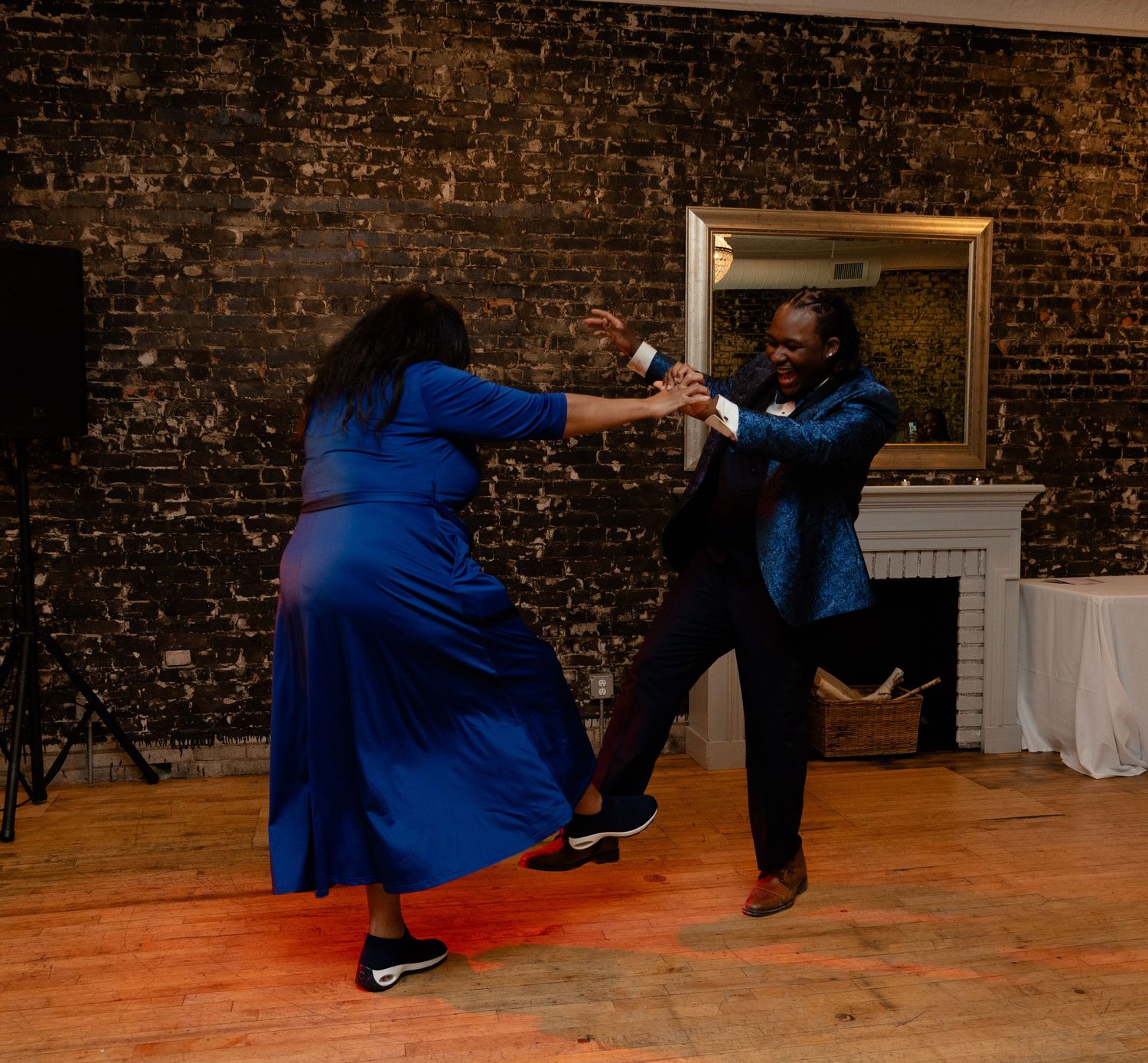 Two dancers in vibrant blue outfits perform an energetic dance move on a wooden floor in front of a brick wall.