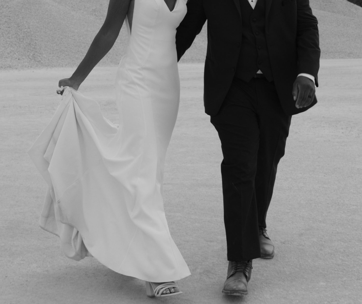 Black and white photo showing the lower half of a bride in white gown walking beside groom in dark suit.
