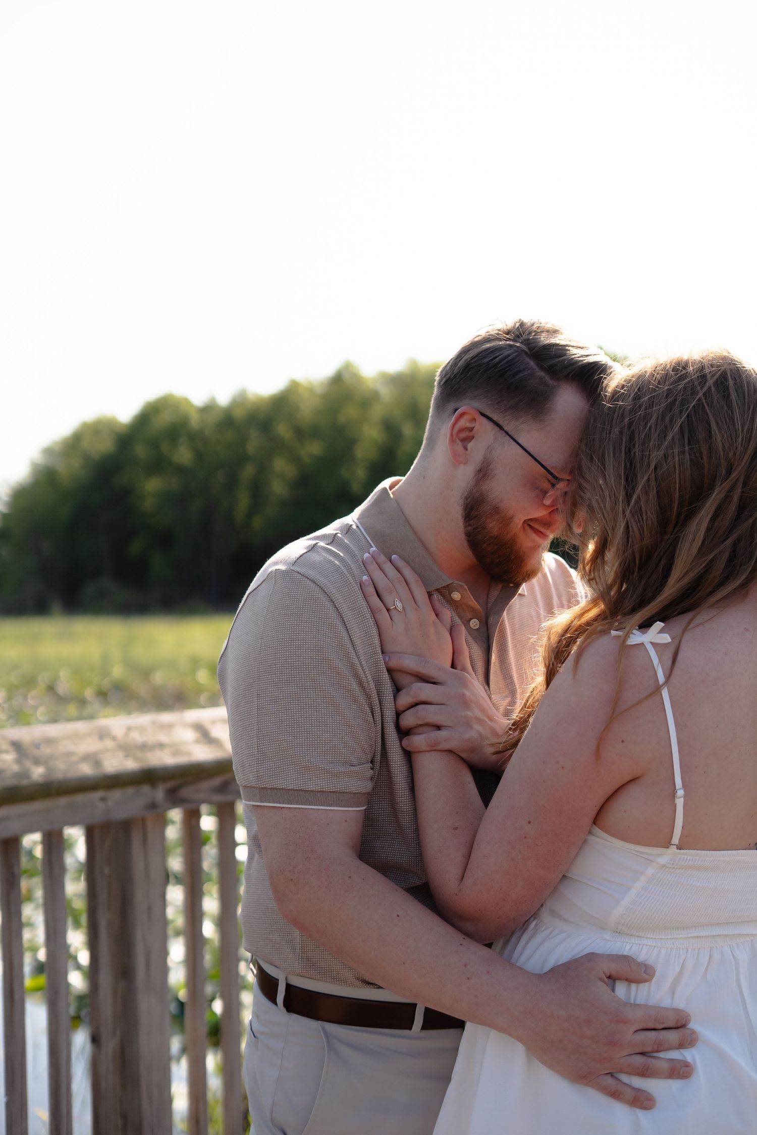 A romantic couple embraces on a wooden deck at sunset, surrounded by green trees and natural scenery.
