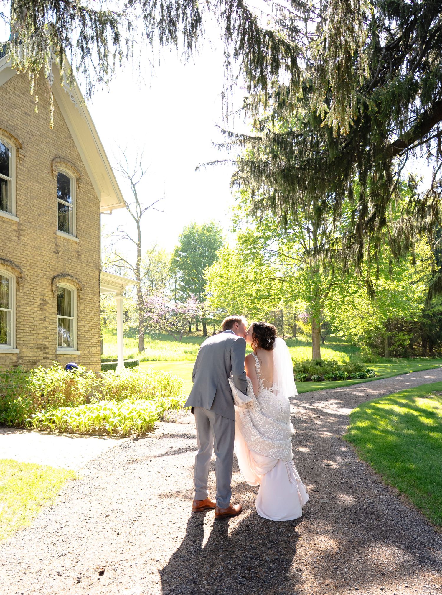A couple in formal wedding attire walking down a gravel path beside a yellow brick building on a sunny spring day.