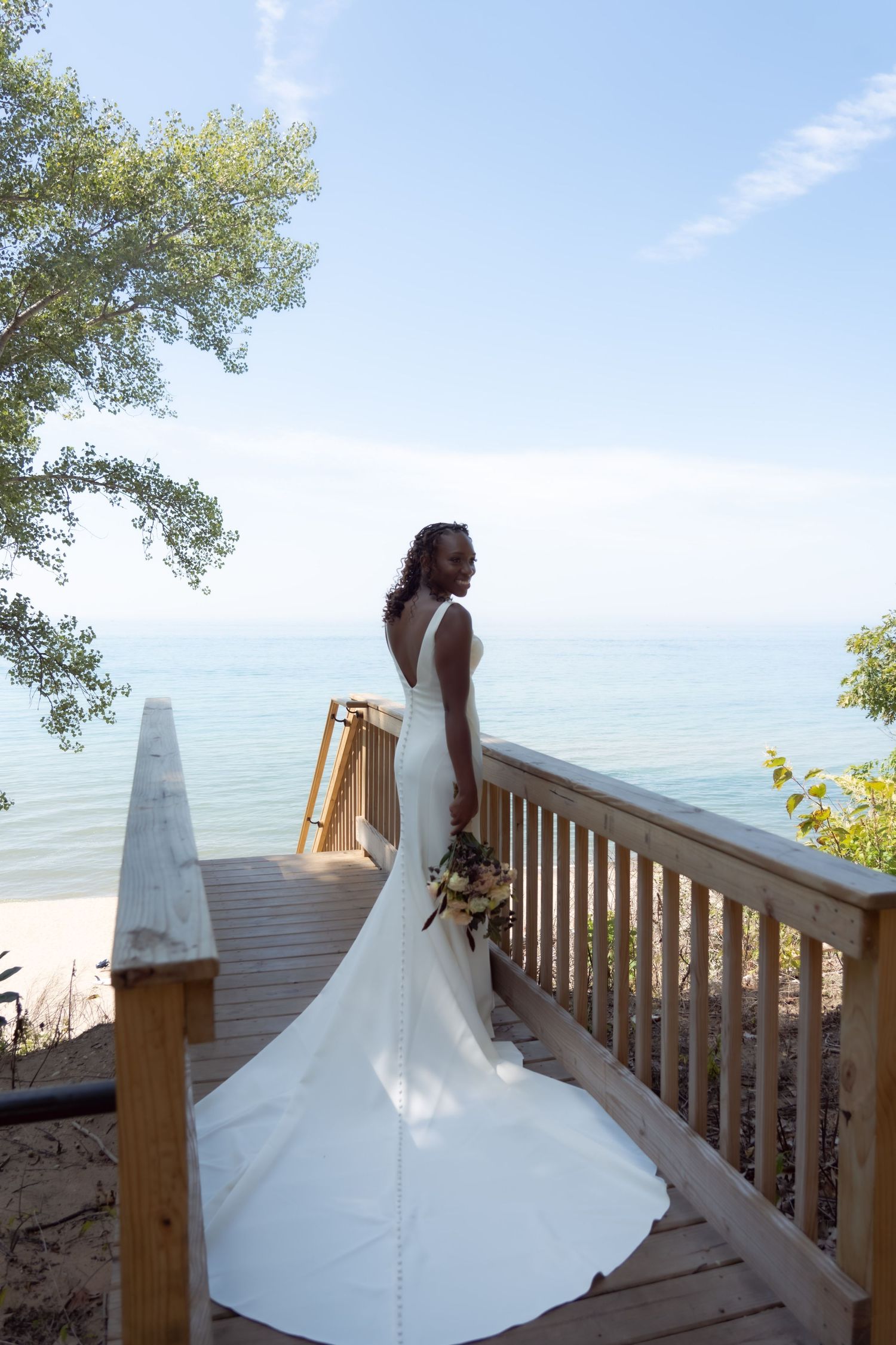 Bride in elegant white dress stands on oceanfront deck with flowing train and seaside view.