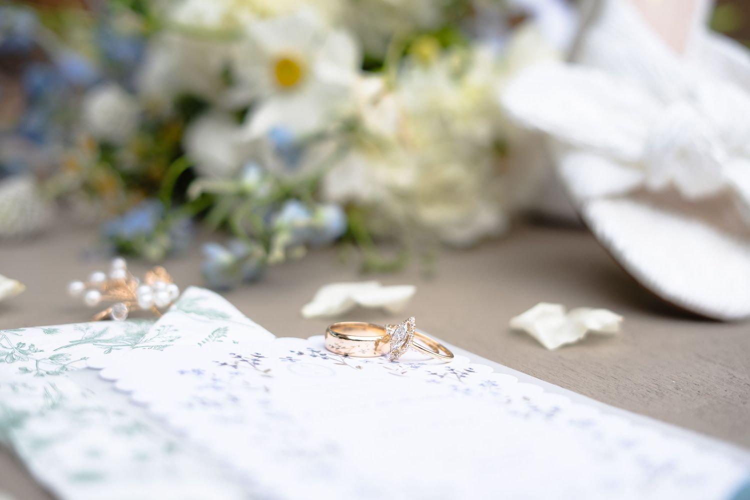 Wedding rings rest on a delicate invitation with soft white flowers and petals scattered in the background.