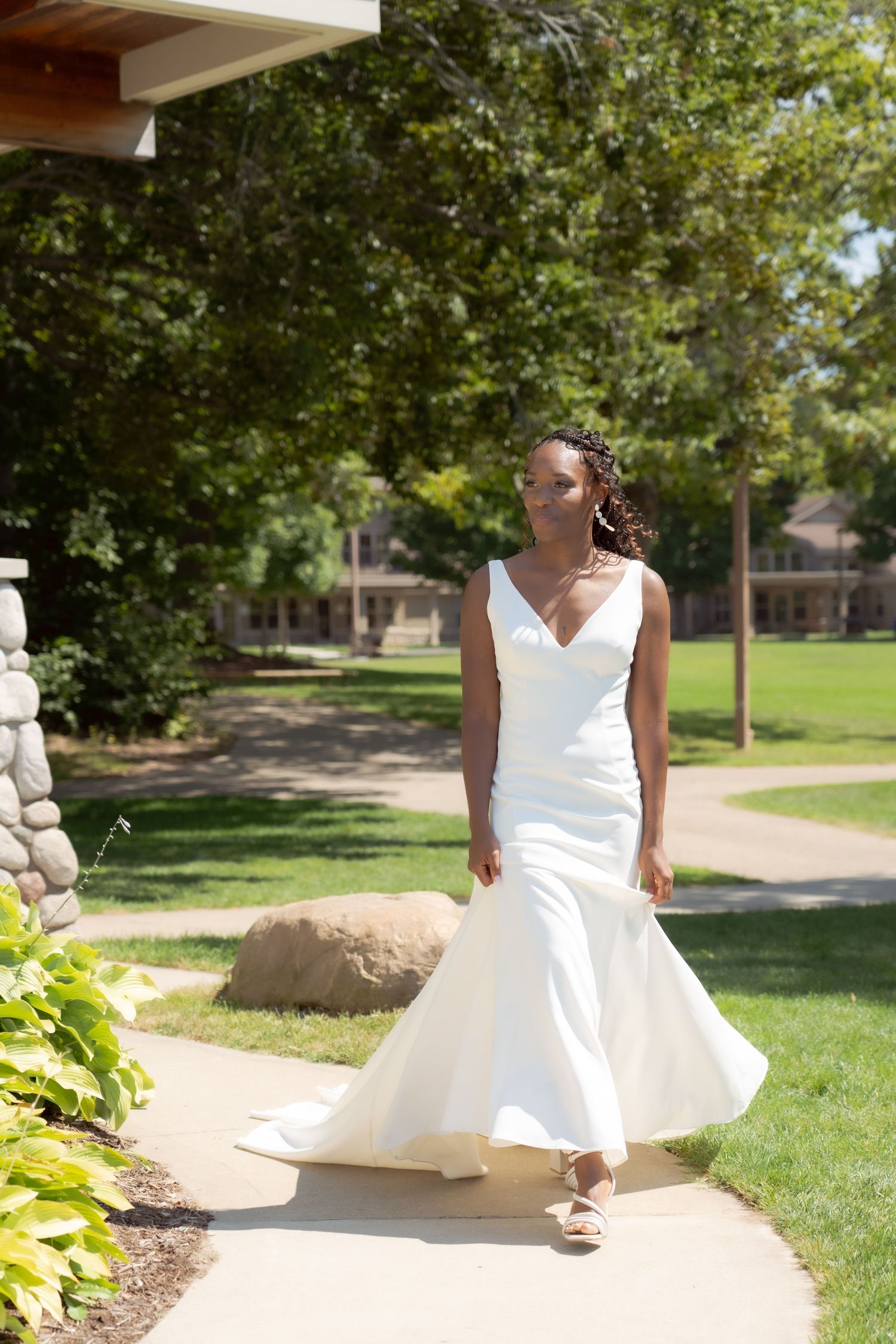 A bride in a flowing white mermaid-style wedding dress walks along a garden path on a sunny summer day.