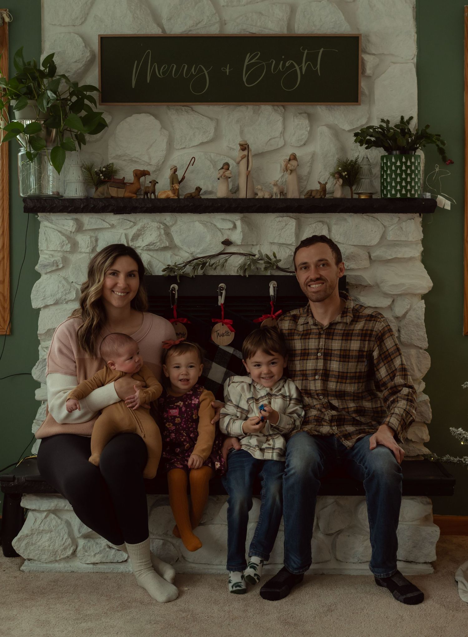 Family holiday photo in front of a stone fireplace with rustic 'Merry + Bright' decor.