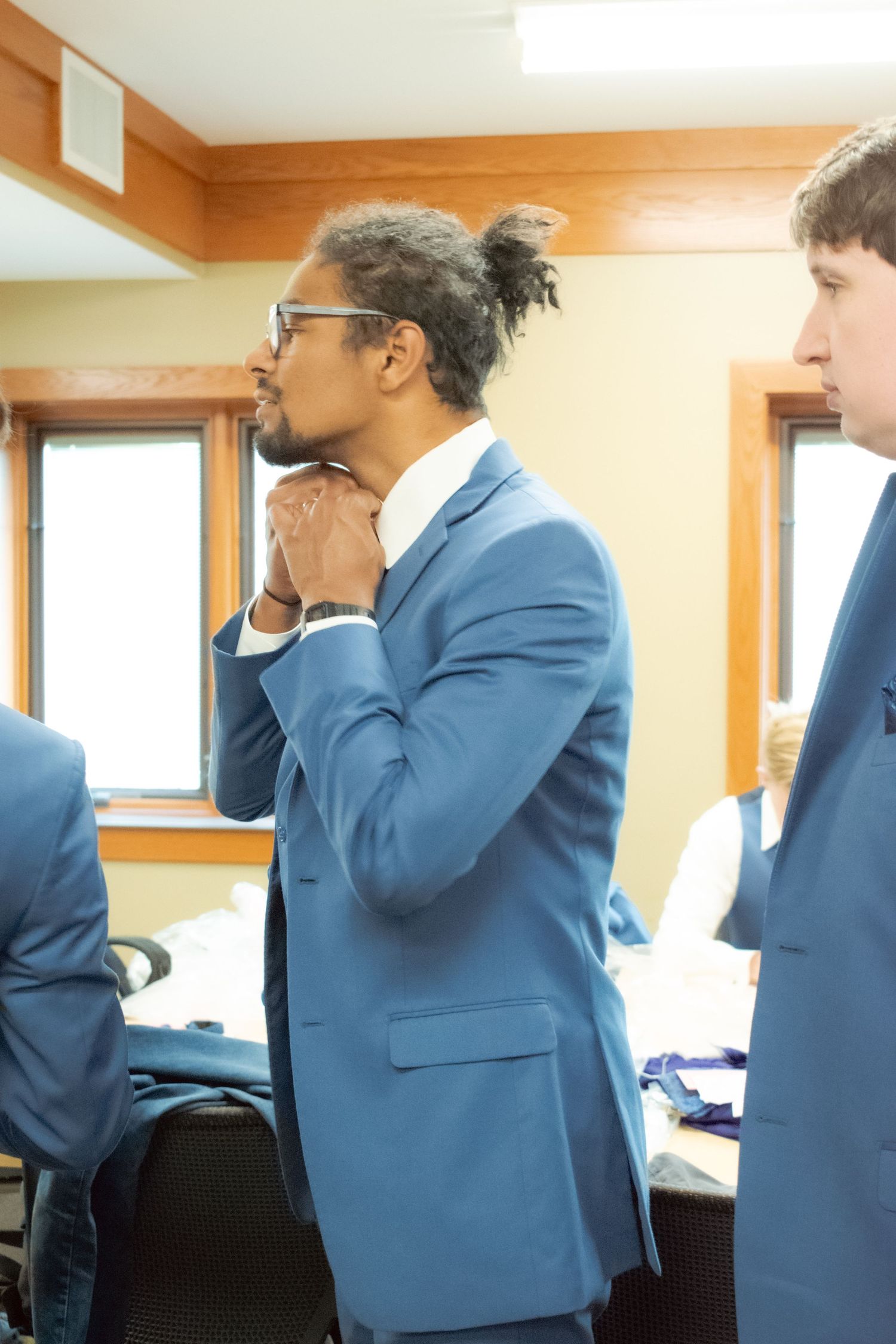 Professional in a light blue suit stands thoughtfully in an office setting with hand on chin.