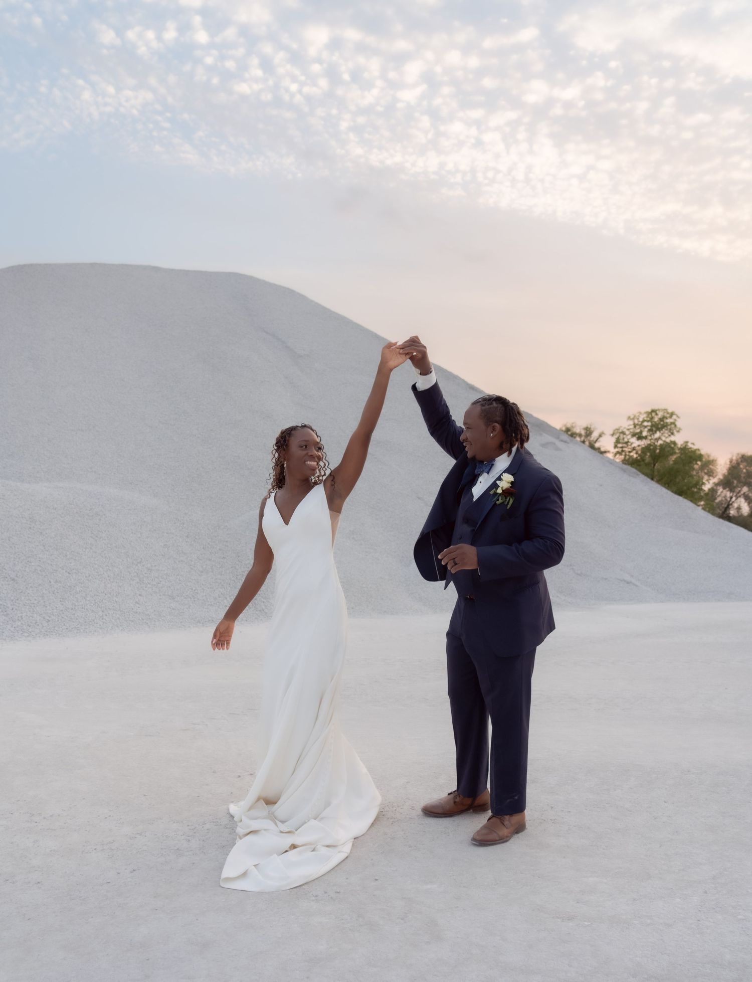 A couple shares a joyful dance moment against a white sandy landscape at sunset.