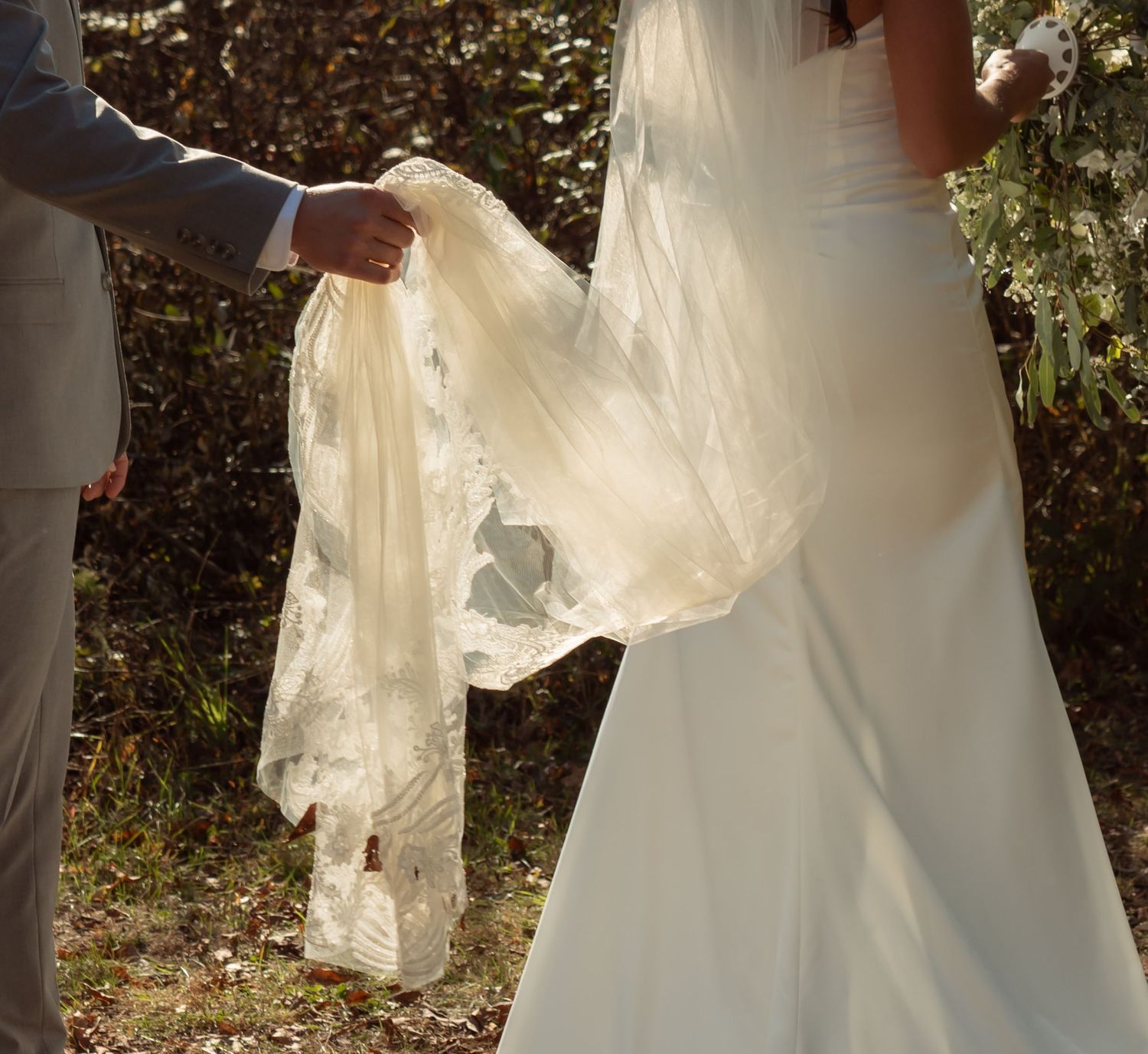 A delicate lace wedding veil is held up in sunlit outdoor setting next to a white wedding dress.