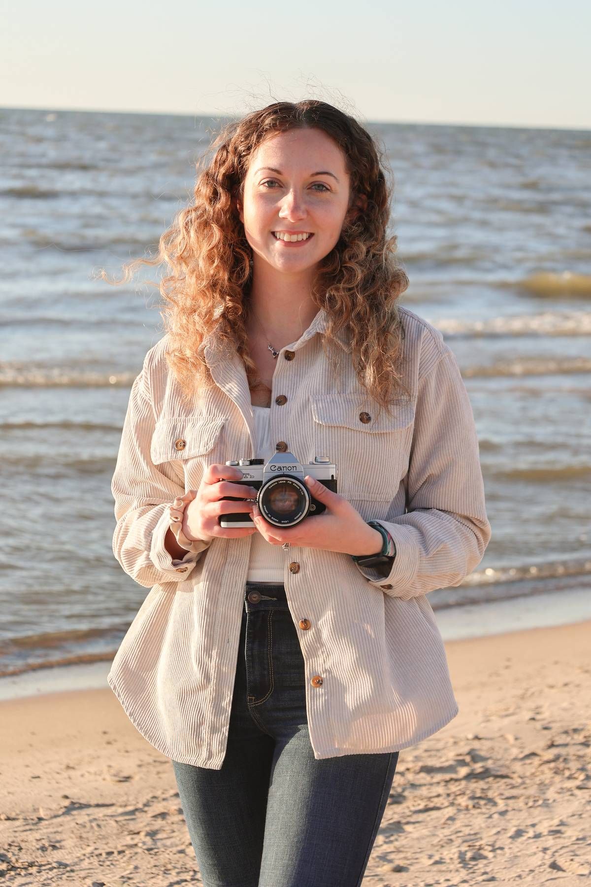A photographer with curly hair holds vintage camera on sunny beach.