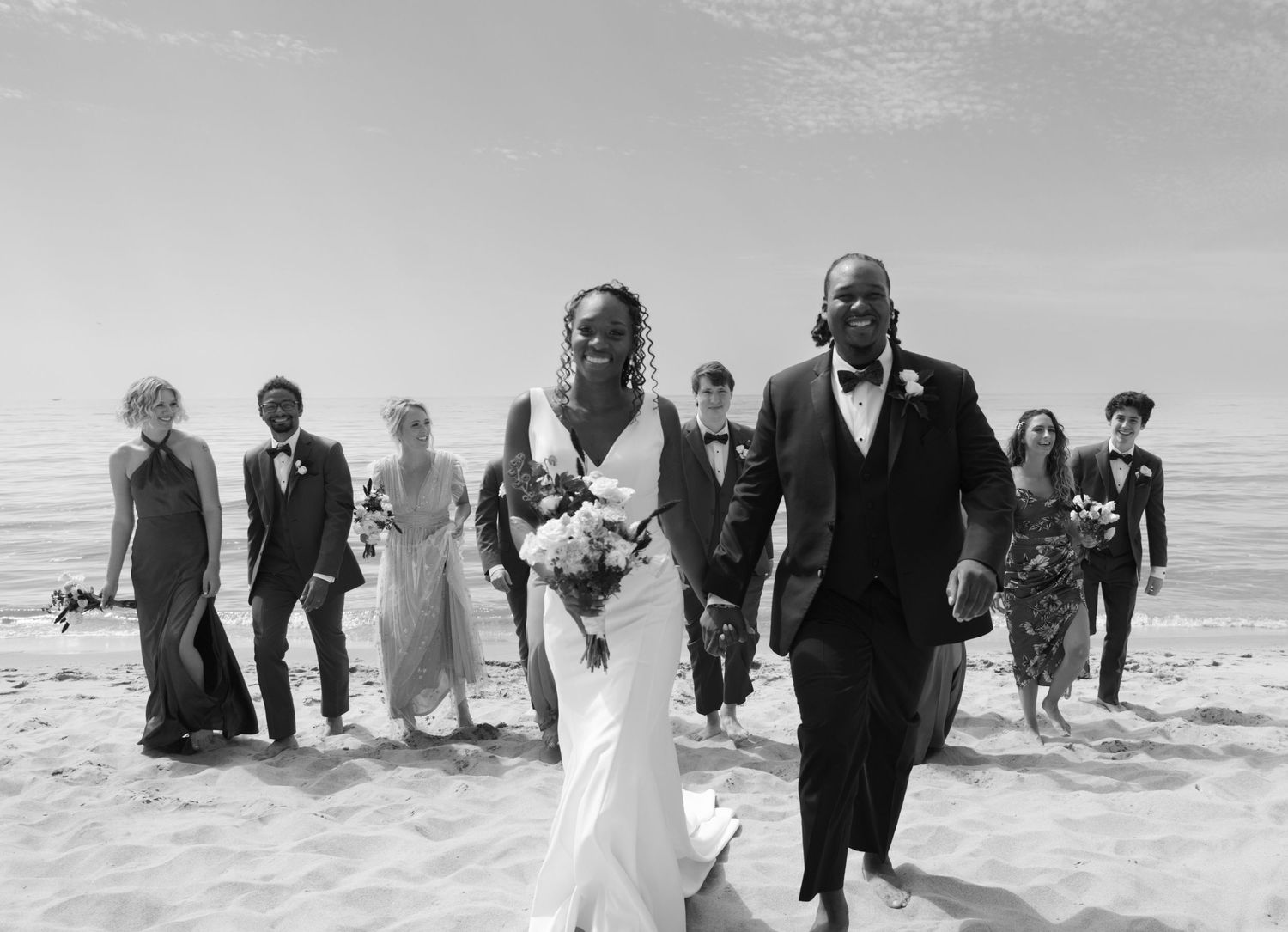 A joyful wedding party walks and laughs together along a sun-lit beach in black and white photography.