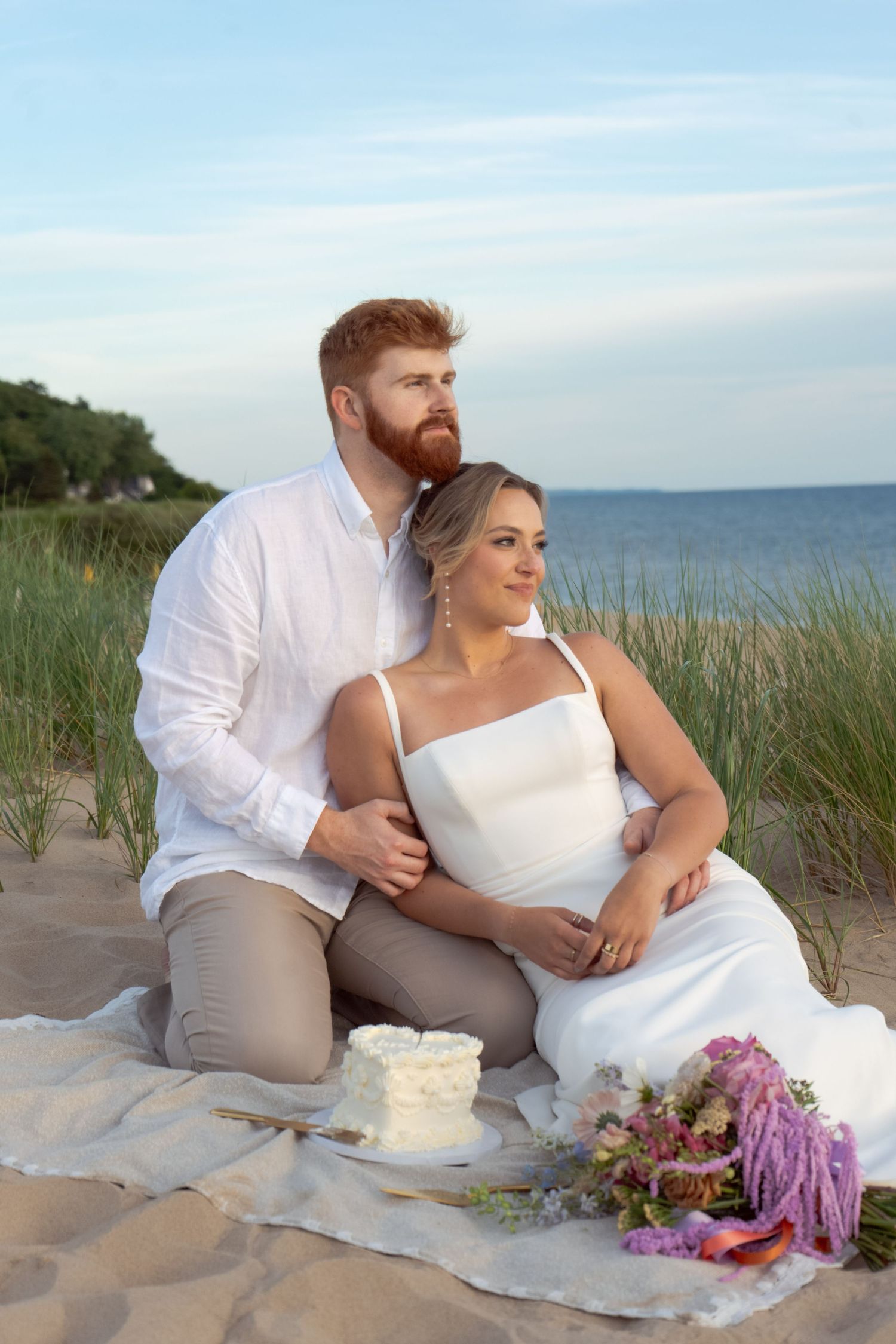 Romantic beach wedding picnic setup with fresh flowers and cake on sand dunes at sunset.
