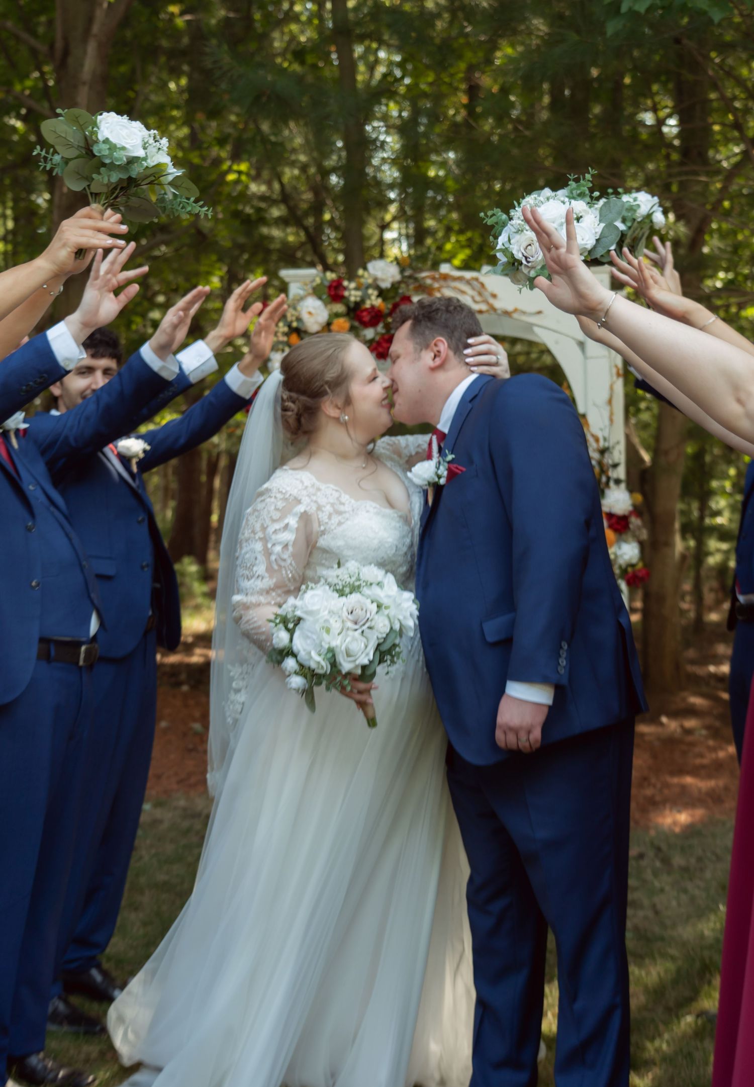 Wedding party holds bouquets high as couple shares first kiss surrounded by trees in outdoor ceremony.